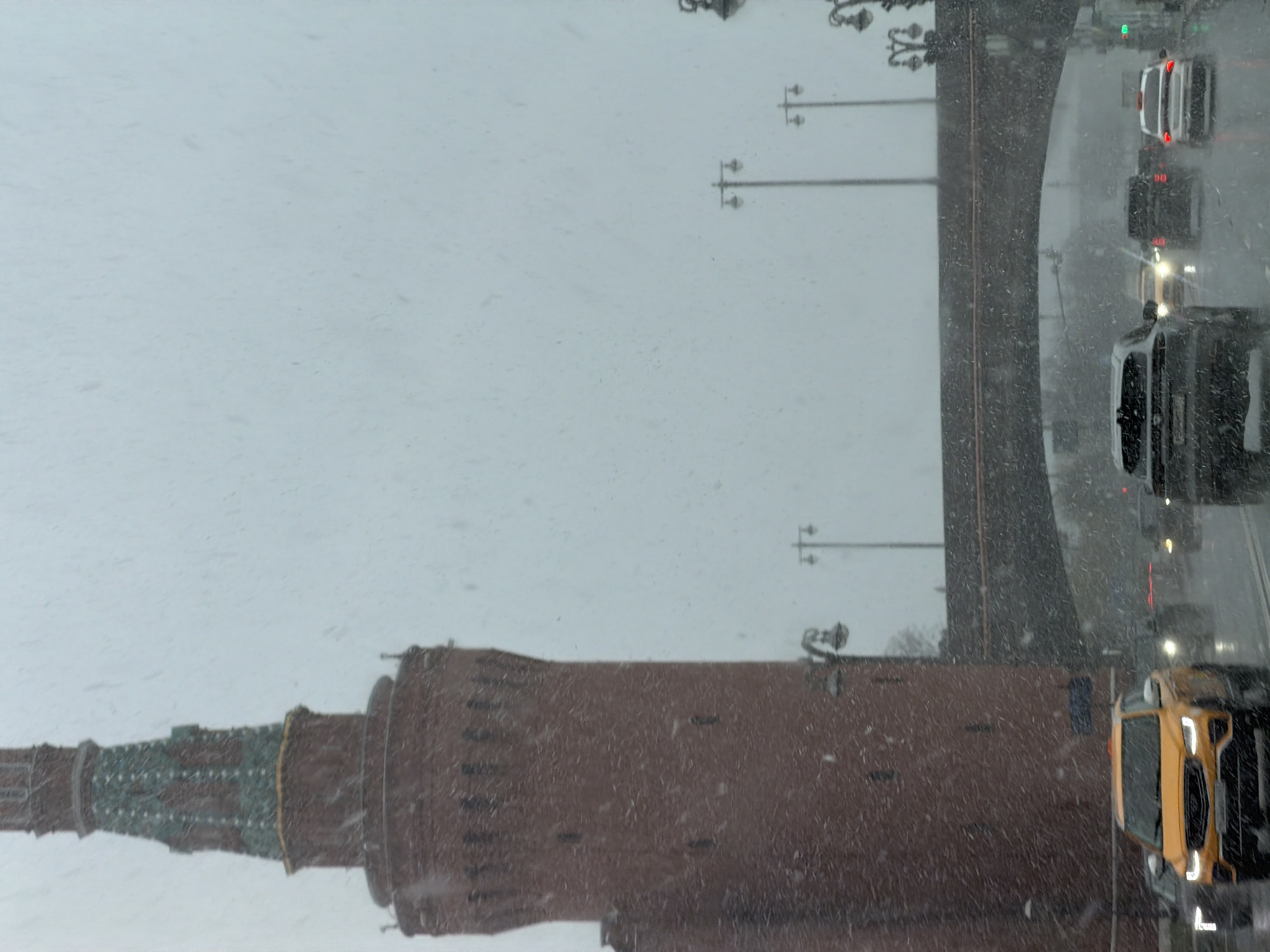 The image depicts a snowy scene in Moscow, Russia. In the foreground is the distinctive brick wall and tower of the Moscow Kremlin. A bridge spans over a road, with several cars visible driving in snowy conditions. Streetlights line the bridge. The sky is overcast and grey. Snowflakes are visible throughout the image.