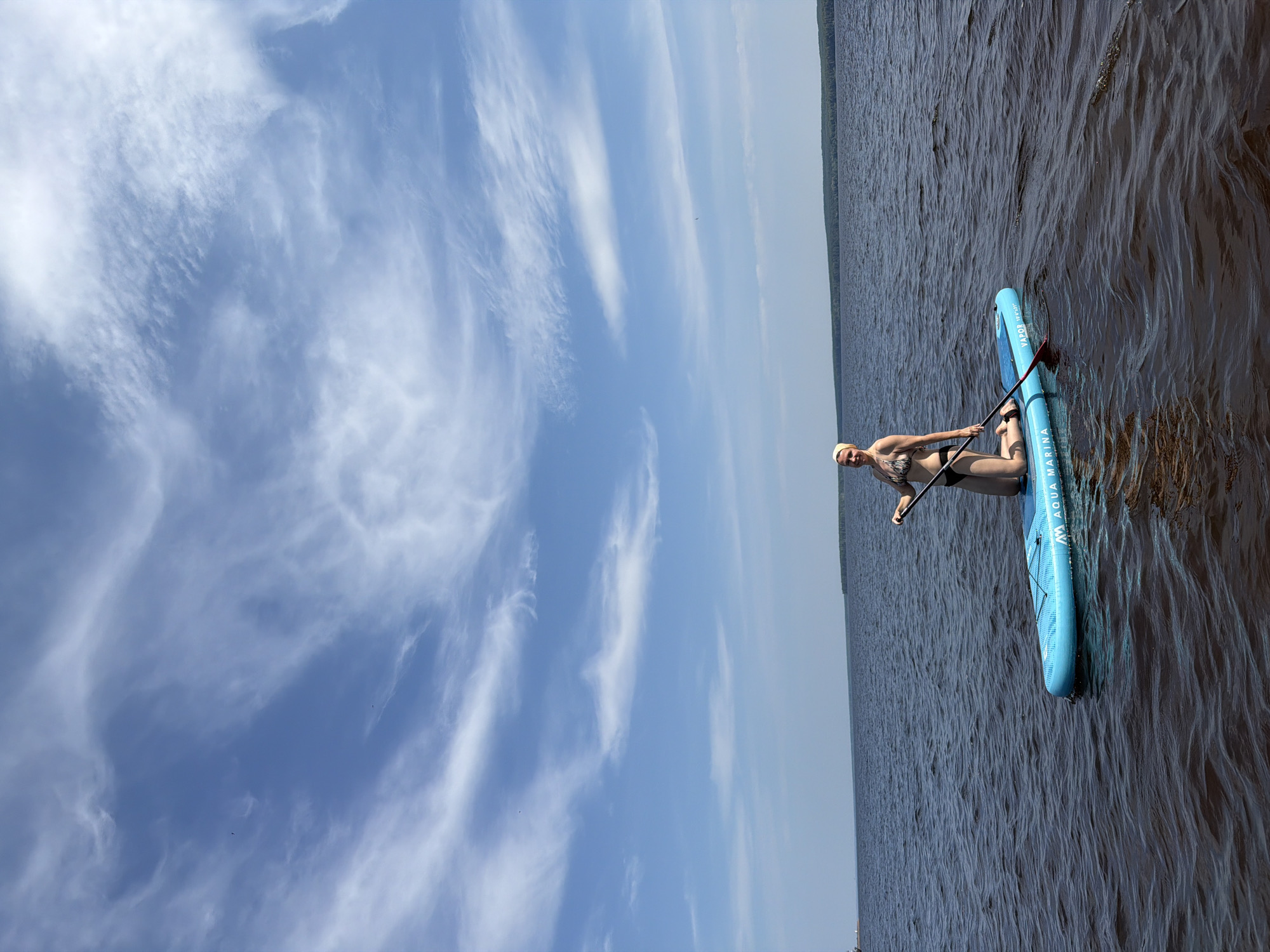 A woman is paddleboarding on a body of water. She is kneeling on a blue paddleboard with a paddle in her hands. She is wearing a bikini and a yellow headscarf. The water is dark and choppy. The sky is blue with wispy white clouds. In the distance, there is a tree line. The location is the Kama River in Chaikovsky, Russia.