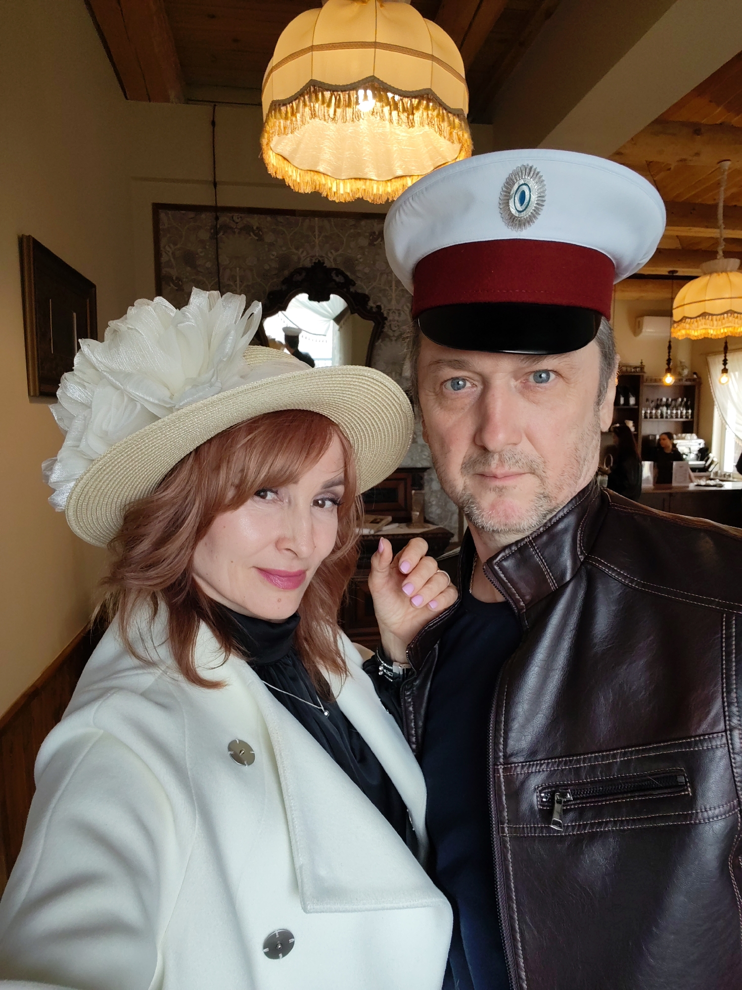 A medium shot depicts a woman and a man in vintage attire, appearing to be in a staged photograph, taken inside a location. The woman, left of the frame, wears a cream-colored hat adorned with a large white feather embellishment, a white overcoat, and a black top with a silver necklace. Her auburn hair is styled with side-swept bangs, and she has makeup on. The man, to the right, dons a tall white cap with a red band and a silver emblem. He is wearing a brown leather jacket over a dark shirt. The light fixtures and mirror in the background give an idea of the ambiance. The photograph was taken in a museum-restaurant, and the location is in the city of Sarapul, Russia.