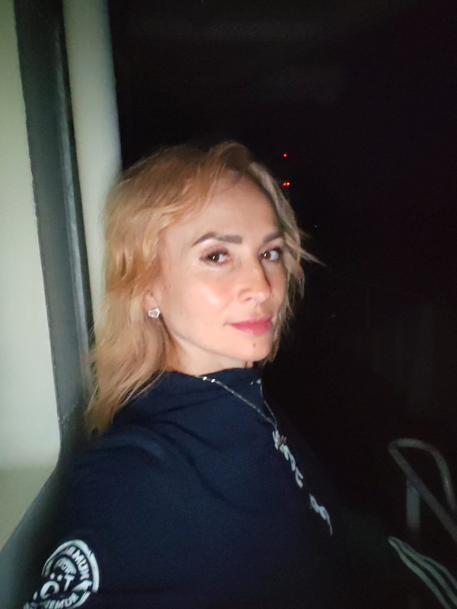 A woman with blonde hair and makeup is photographed indoors, leaning against a white vertical surface. She is wearing a dark hooded jacket with a silver zipper and a pendant necklace. Her right ear is adorned with a heart-shaped earring. The background is dark, with some blurry red lights visible in the distance, suggesting an evening or night setting. A portion of a black railing is visible in the lower right corner. The scene appears to be inside a train or bus, given the narrow, enclosed space and the presence of a railing. The location is not specified as Ivanov Bor, Russia, and no such information can be inferred from the image itself.