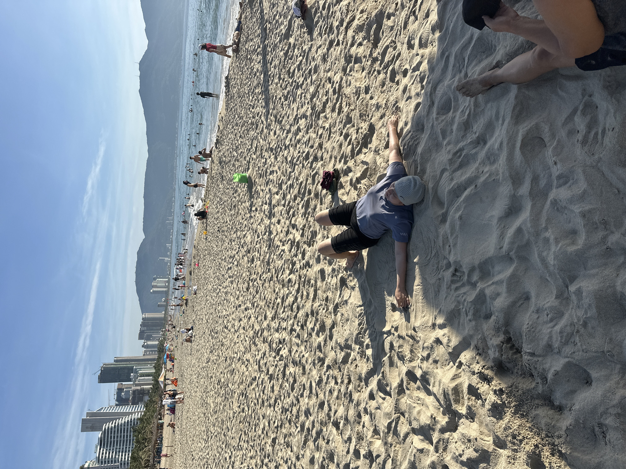 The image depicts a crowded beach scene in Da Nang, Vietnam, likely My Khe Beach, with a backdrop of modern high-rise buildings and distant mountains. In the foreground, a person lies supine on the sand, arms outstretched, wearing a grey t-shirt, dark shorts, and a grey beanie. The sand is textured with numerous footprints and small undulations. Further into the scene, other beachgoers are visible, some walking along the shore, others in the water, and some relaxing on the sand. A green bucket stands near the water's edge. The ocean appears calm with small waves rolling in. The sky is clear with a few wispy clouds, and the sun is casting shadows across the sand, indicating it is likely late afternoon.