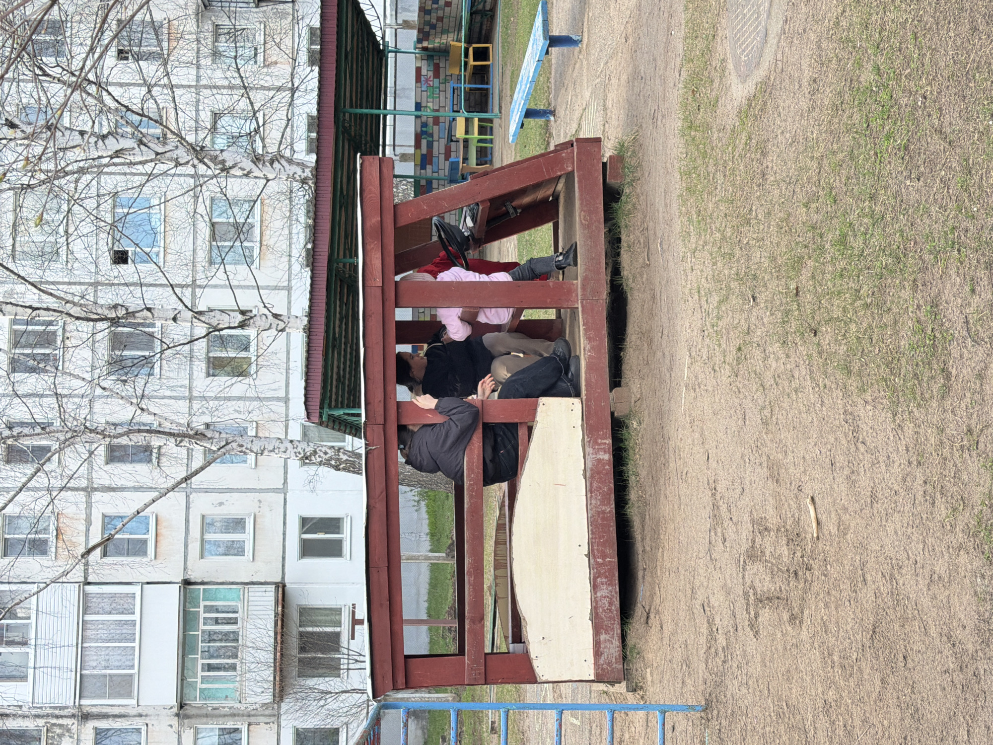 The image depicts a children's playground in Chaikovsky, Russia. Three people are inside a red-brown wooden structure. One is wearing a black jacket, another a pink shirt, and the third is in black and beige clothing. An apartment building is in the background, partially obscured by bare branches. The ground is covered in sand and sparse grass. A blue bench and a blue playground structure are also visible.