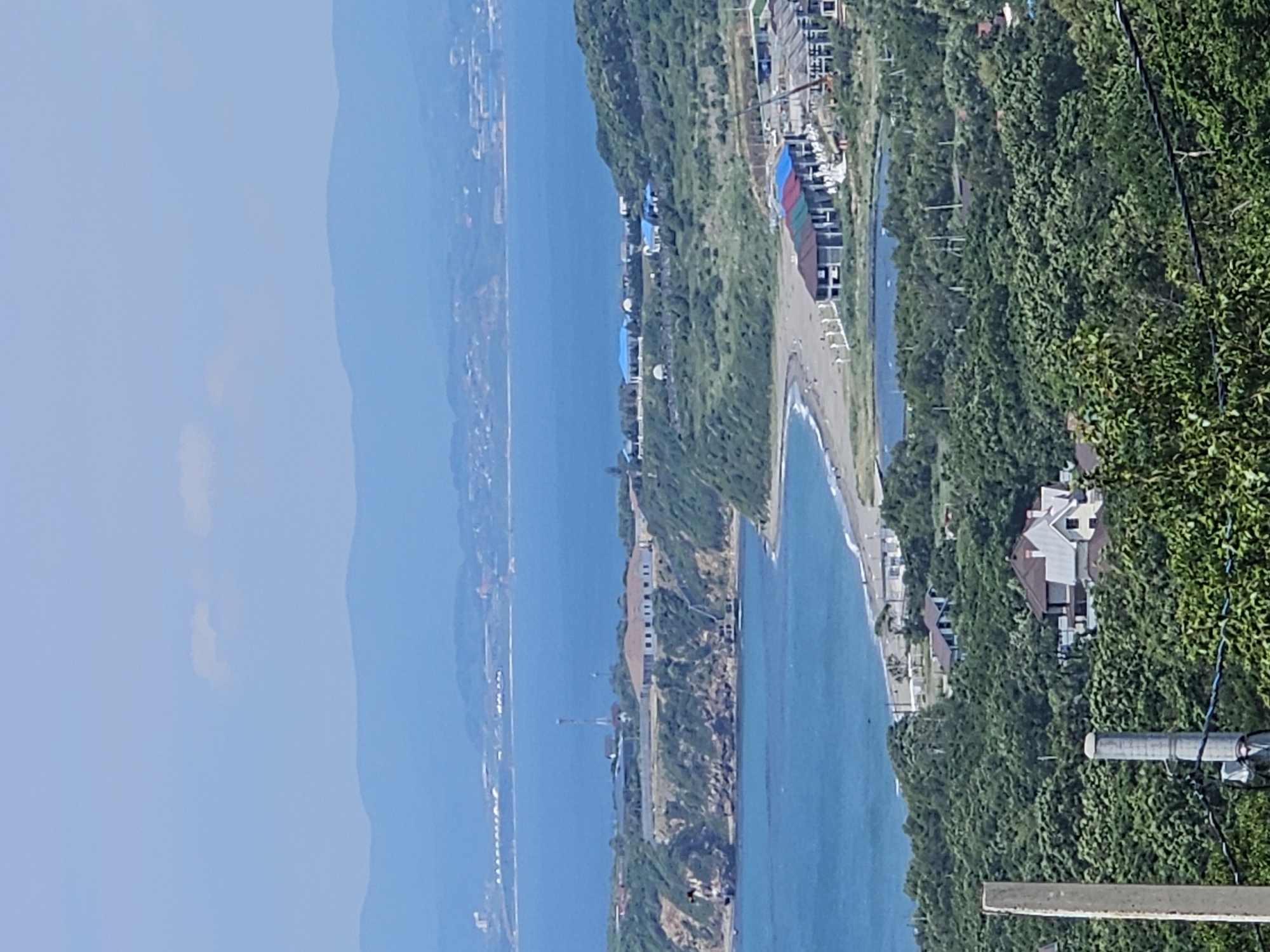 The image is a high-angle, panoramic view of a coastal area in Nakhodka, Russia, on a clear, sunny day. In the foreground, dense green foliage of trees and shrubs obscures the lower portion of the scene, with two light-colored, cylindrical posts partially visible. Beyond the foliage, a cluster of houses with dark roofs, including one light-colored house with a dark roof and several dark-roofed houses, are nestled amongst the trees.
Further back, a body of water, possibly a pond or small lake, is situated in a clearing within the trees. Adjacent to this water body, a complex of buildings under construction is visible, featuring colorful roofs (red, green, and blue) and visible construction cranes.
The mid-ground reveals a sandy beach stretching along the coastline, with a few indistinct figures walking on the sand. The sea, a vibrant blue, meets the shore. Along the coast, on a grassy, elevated bluff, a long, white building with a dark roof is prominent, with a tall radio tower nearby. The bluff is covered in green vegetation and features some rocky outcrops.
In the background, the calm sea extends towards a distant coastline with layered mountains fading into the hazy blue sky. A thin strip of structures, possibly a city or port, can be seen along the distant shoreline. The sky is light blue with a few faint, white clouds.