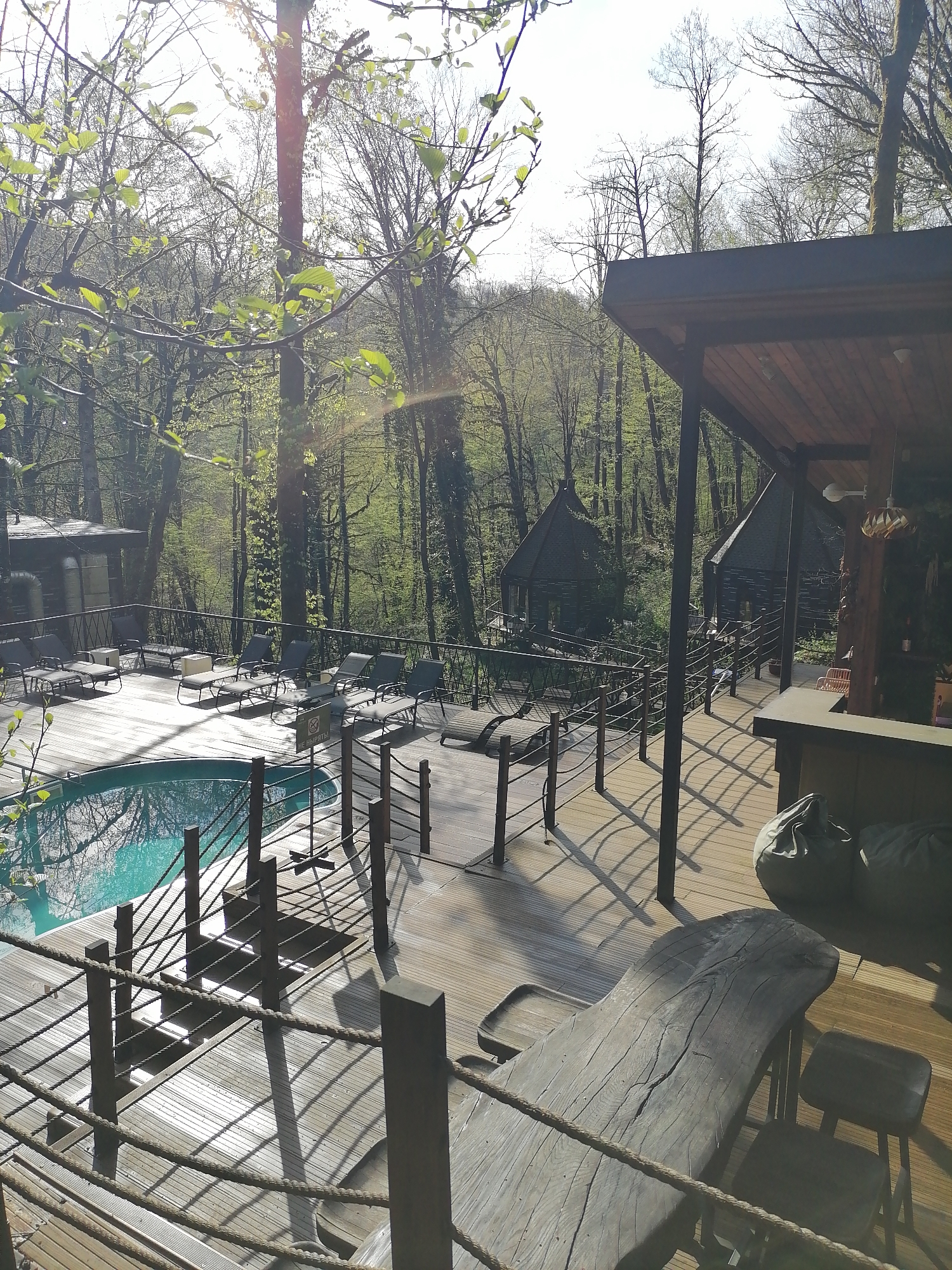 The image displays an outdoor leisure area, likely part of a resort or glamping site, nestled within a dense forest. A spacious wooden deck forms the main platform, featuring a rounded swimming pool with clear, light blue water that reflects the surrounding trees. Multiple modern lounge chairs are arranged around the pool, suggesting a sunbathing and relaxation zone.
On the right side of the deck, under a wooden patio cover, a large, rustic wooden table with a natural edge design is visible, accompanied by stools or low chairs. Two sizeable grey beanbag cushions rest near this seating arrangement. The deck is secured by a distinctive rope railing system with wooden posts, casting long, sharp shadows across the wooden planks.
In the midground and background, several unique dark-colored structures resembling cabins or bungalows are situated among the tall trees, which are mostly bare-branched but show some early spring foliage. Some of these structures feature multi-faceted or conical roofs. A larger, more conventional building is visible further back on the left side of the deck.
The sun is low in the sky, creating prominent shadows and a strong lens flare in the upper left corner, indicating either early morning or late afternoon. The weather is clear and bright. No people are present, lending a serene and secluded atmosphere to the scene. A small white sign positioned near the pool clearly reads "НЕ НЫРЯТЬ!", which translates from Russian as "DO NOT DIVE!".