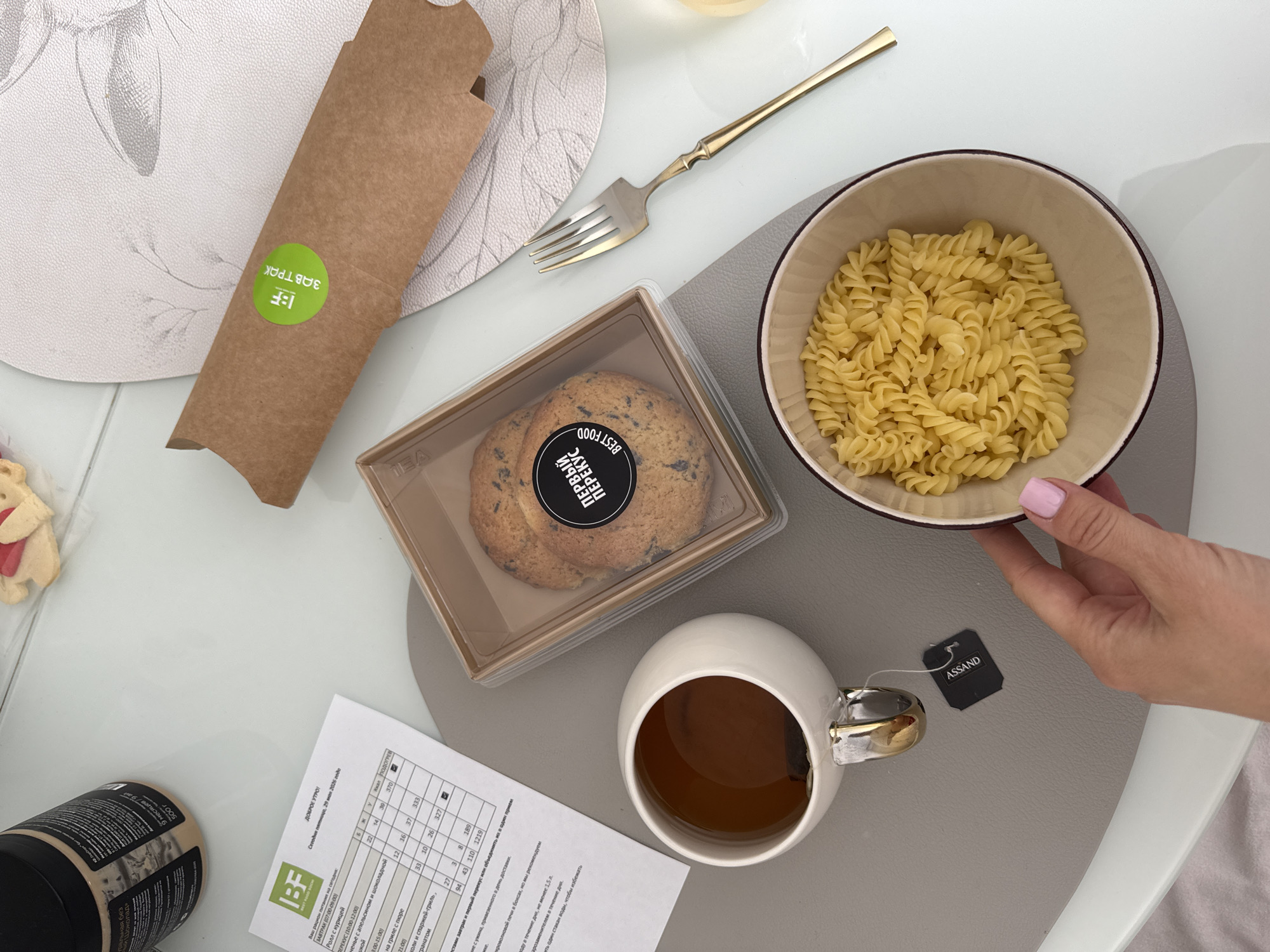 A top-down shot captures a light-filled scene on a table, likely indoors, with various food items arranged. A person's hand with pink nail polish holds a bowl of cooked pasta. To the left of the bowl, there's a white mug of tea with an "ASSAND" tea bag tag. Below the mug, a printed paper displays nutritional information.
Next to the mug is a transparent container holding two cookies. A brown paper package with a green "1BF ЗАВТРАК" sticker sits diagonally on a placemat. A gold fork is positioned between the pasta bowl and the brown package.
The presence of Russian text on the food packaging and nutritional information suggests this scene is taking place in Russia. The text "ДОБРОЕ УТРО! Сегодня пятница, 29 мая 2026 года" indicates it's a Friday morning on May 29, 2026. Based on the provided information on the nutritional paper, the food items seem to come from a local restaurant. The information on the menu "ЗАВТРАК (07:00-09:00)" with options "Ролл с курицей", and location info on the paper, suggests the city is Sochi, Russia.
