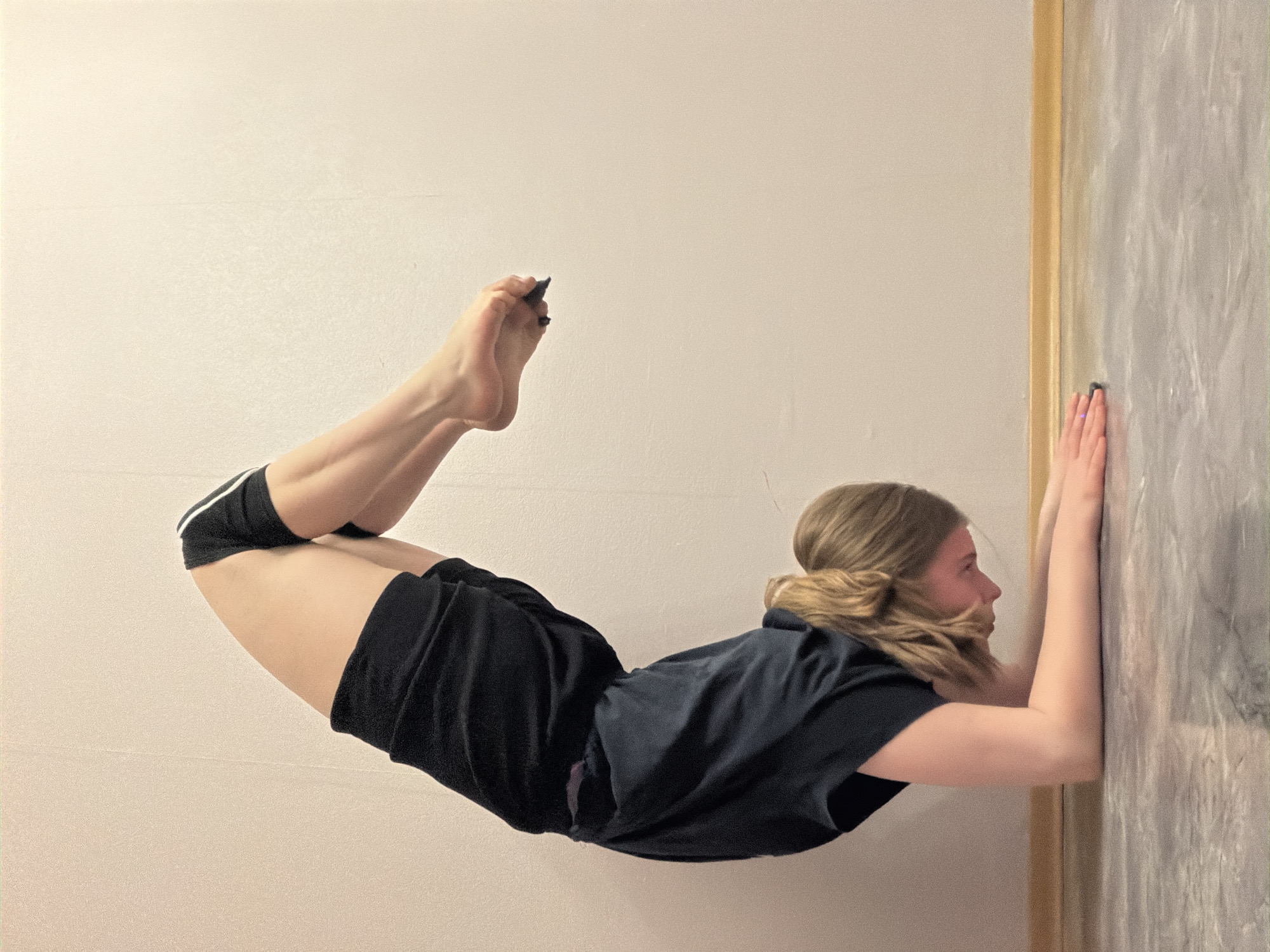 A young woman is performing a handstand against a wall. She is wearing black shorts and a black T-shirt. Her hair is blonde and pulled back in a bun. She is positioned parallel to the camera and the wall, with her hands placed flat against the wall at about shoulder height. Her body is extended outward, legs bent at the knee and extended upward. The background consists of two walls of different textures, the one on the left, with a smooth beige-pink color. The one on the right appears to be a textured painted wall, possibly with a frame around it. The location is likely indoors, but the exact place cannot be identified in Perm, Russia based on the image.