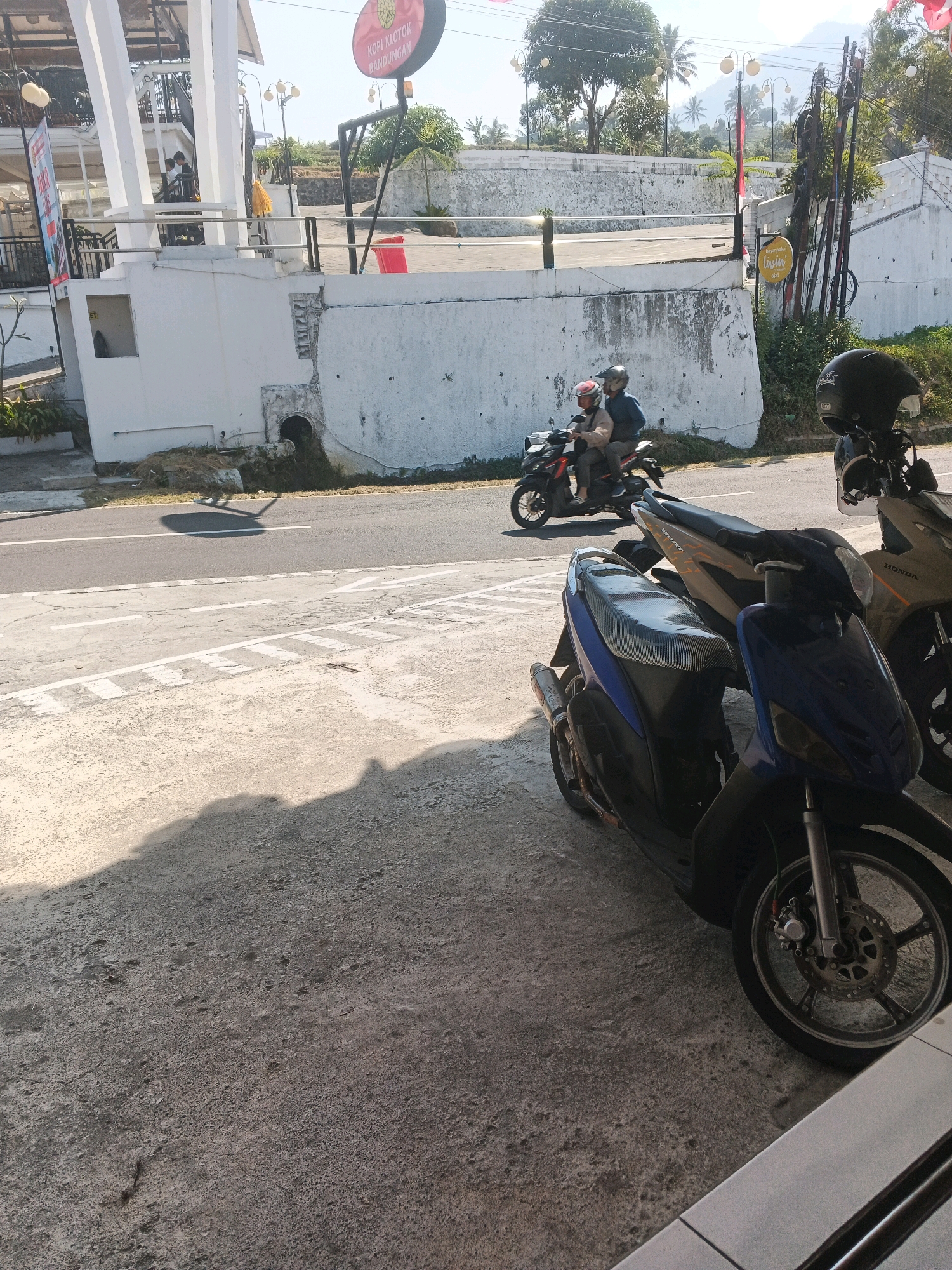 The image captures a street scene in Pakopen, Indonesia, on a sunny day. In the foreground, two parked motorcycles are visible, one blue and one tan. The blue motorcycle is closer to the viewer and is a scooter with a striped seat cover. The tan motorcycle is behind it. Further down the street, a red motorcycle is ridden by two individuals wearing helmets. The rider in the back is a person wearing a blue jacket, and the person in front is wearing a light-colored jacket. Behind the motorcycles, a white wall marks the boundary of a property, with some vegetation growing around it. Across the street, a white building with pillars is visible, and a circular sign with red and yellow lettering hangs from a pole. The sky is clear and blue, indicating fair weather. The overall impression is of a typical street in a town or city, with everyday activities taking place.
