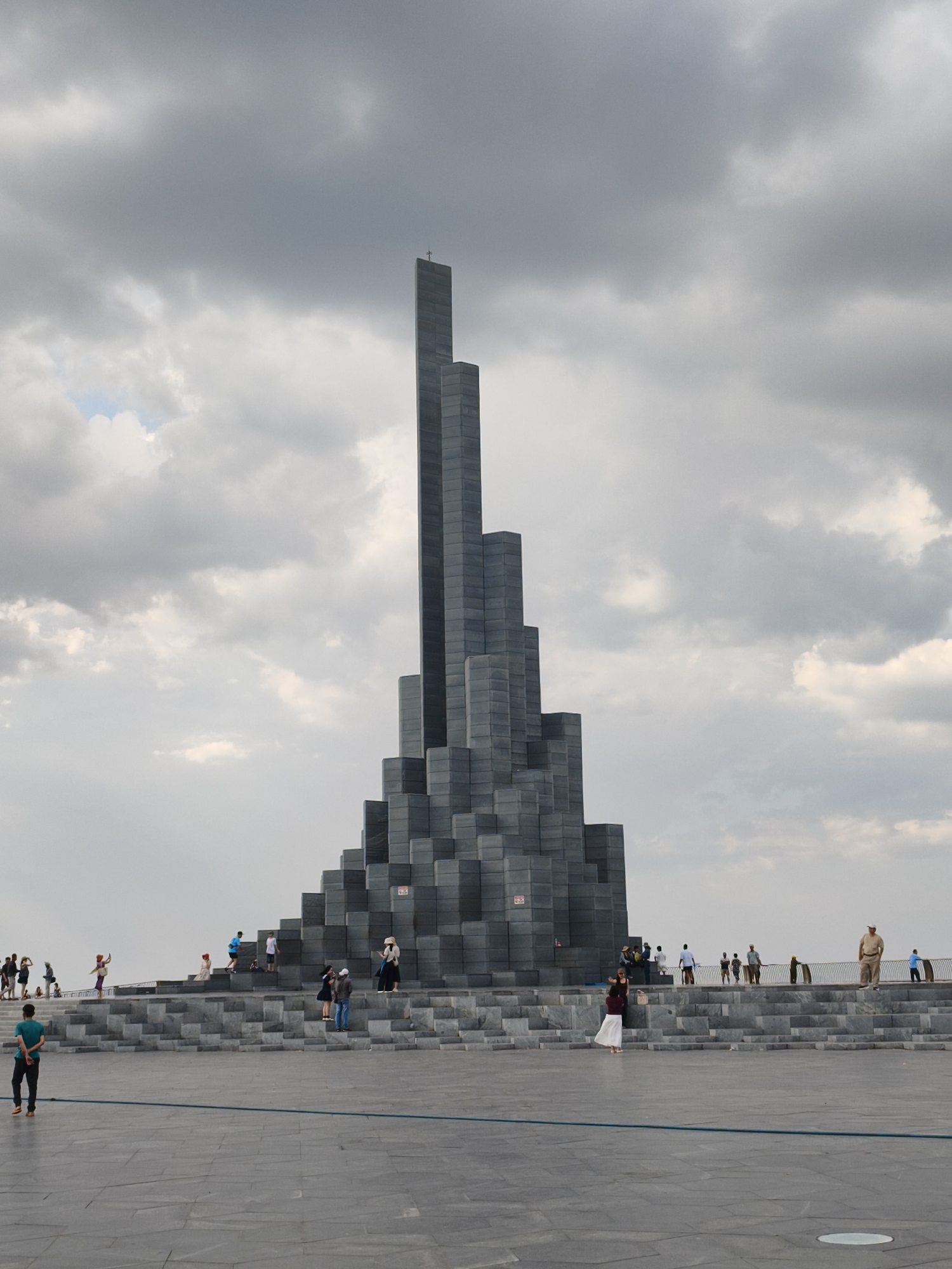 The image shows a large, modern monument composed of dark, angular blocks, resembling stacked geometric shapes. A cross is visible at the apex of the tallest section. The monument is surrounded by broad, paved plazas and tiered steps, also constructed from stone. Several individuals are scattered across the plaza and on the steps, some observing the monument, others interacting or posing for photographs. In the foreground, a lone man in a teal shirt stands facing away from the camera. The sky is overcast with dramatic, grey clouds. This monument is located at the Nghinh Phong Cape in the city of Tuy Hoa, Vietnam, a coastal promontory known for its scenic views and modern architectural landmarks.