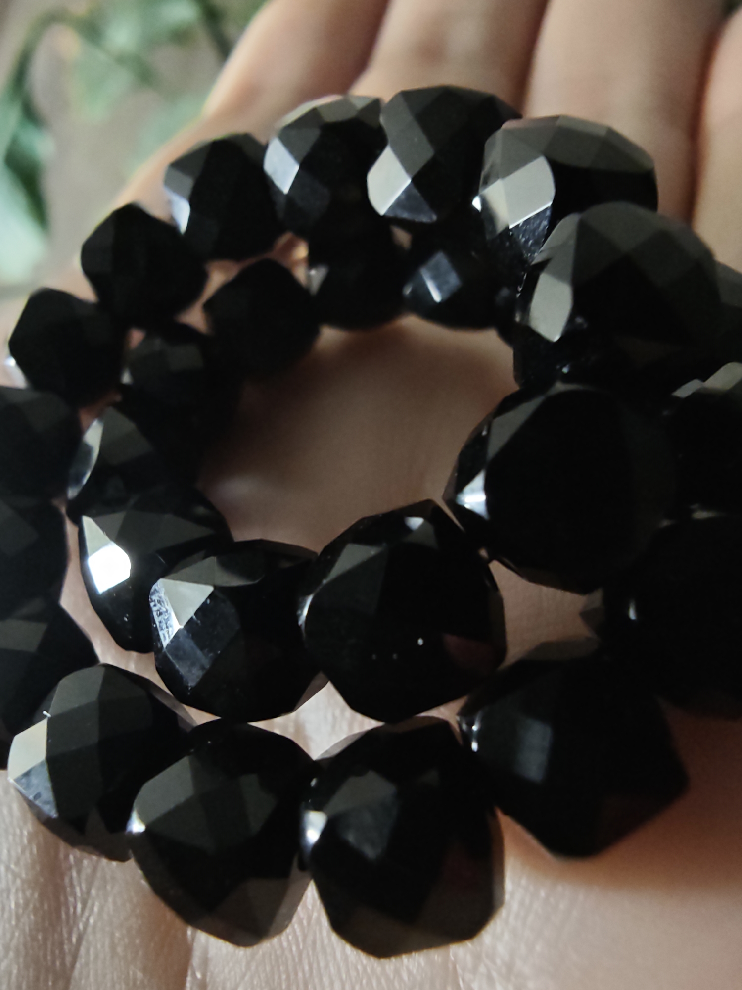 A close-up, slightly angled shot shows a hand holding a black beaded bracelet. The beads are faceted and appear to be made of a dark, reflective material like onyx or obsidian. The hand is positioned to cradle the bracelet, with fingers slightly curled. The background is softly blurred, suggesting an indoor setting with diffused lighting. No specific location in Kursk, Russia, is identifiable from the visual information provided.