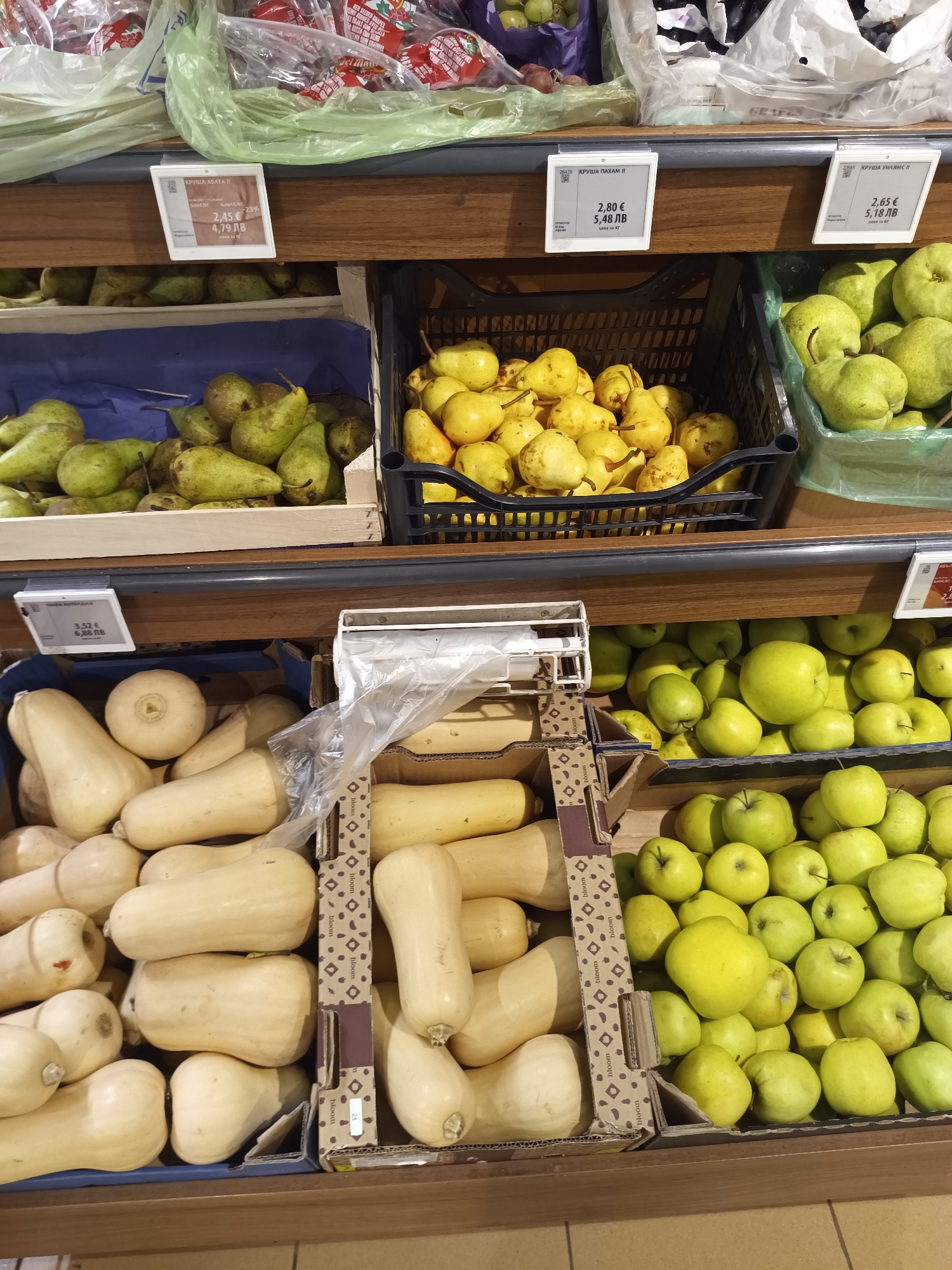 The image shows a supermarket shelf with produce on display. On the top shelf, from left to right, there is a crate of green pears labeled "КРУША АБАТА", a black basket filled with yellow pears labeled "КРУША ПАХАМ II", and a green basket filled with green pears labeled "КРУША УИЛЯМС И". The labels indicate the prices in both euros and Bulgarian leva. On the bottom shelf, there are butternut squash in boxes, and green apples filling out the right side. The boxes of squash have the word "bloom" printed on the sides. There are price labels for the squash as well, but they are partially out of frame. The location is a supermarket, likely in Targovishte, Bulgaria, based on the currency (Bulgarian Leva) on the labels.