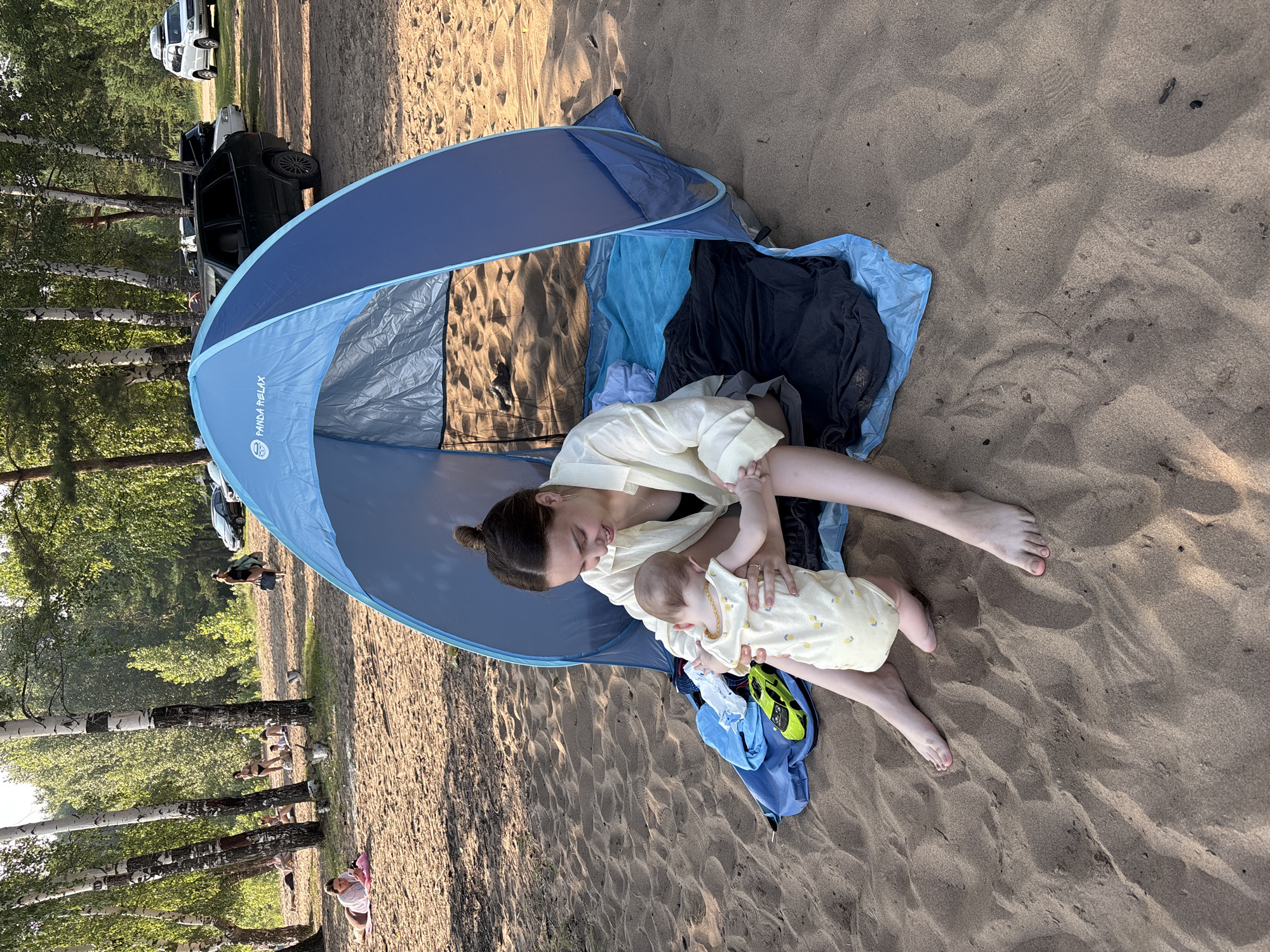 A woman and a baby are sitting on the sand in front of a blue pop-up tent on a beach. The woman is wearing a light-colored button-down shirt and a black tank top, and the baby is wearing a yellow onesie with a lemon pattern. The tent is set up on the sand, with blankets and clothes inside. In the background, there are trees, other people on the beach, and cars parked nearby. The overall scene suggests a relaxed day at the beach. The location is identified as Noviy, Russia.