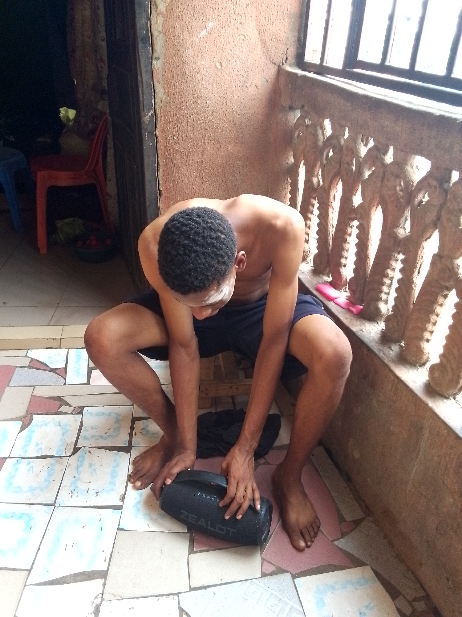A barefoot, dark-skinned man with short black hair is crouching on a tiled floor, interacting with a black speaker marked "ZEALOT." He is wearing dark shorts and is shirtless. The location appears to be an outdoor or semi-outdoor area, with a window or balcony featuring ornate concrete posts. The wall behind the man is rough and painted in a reddish-brown hue. To the left, a doorway leads into a darker interior with chairs and what appears to be produce in a bowl. The location is in the city of Unknown, Unknown.