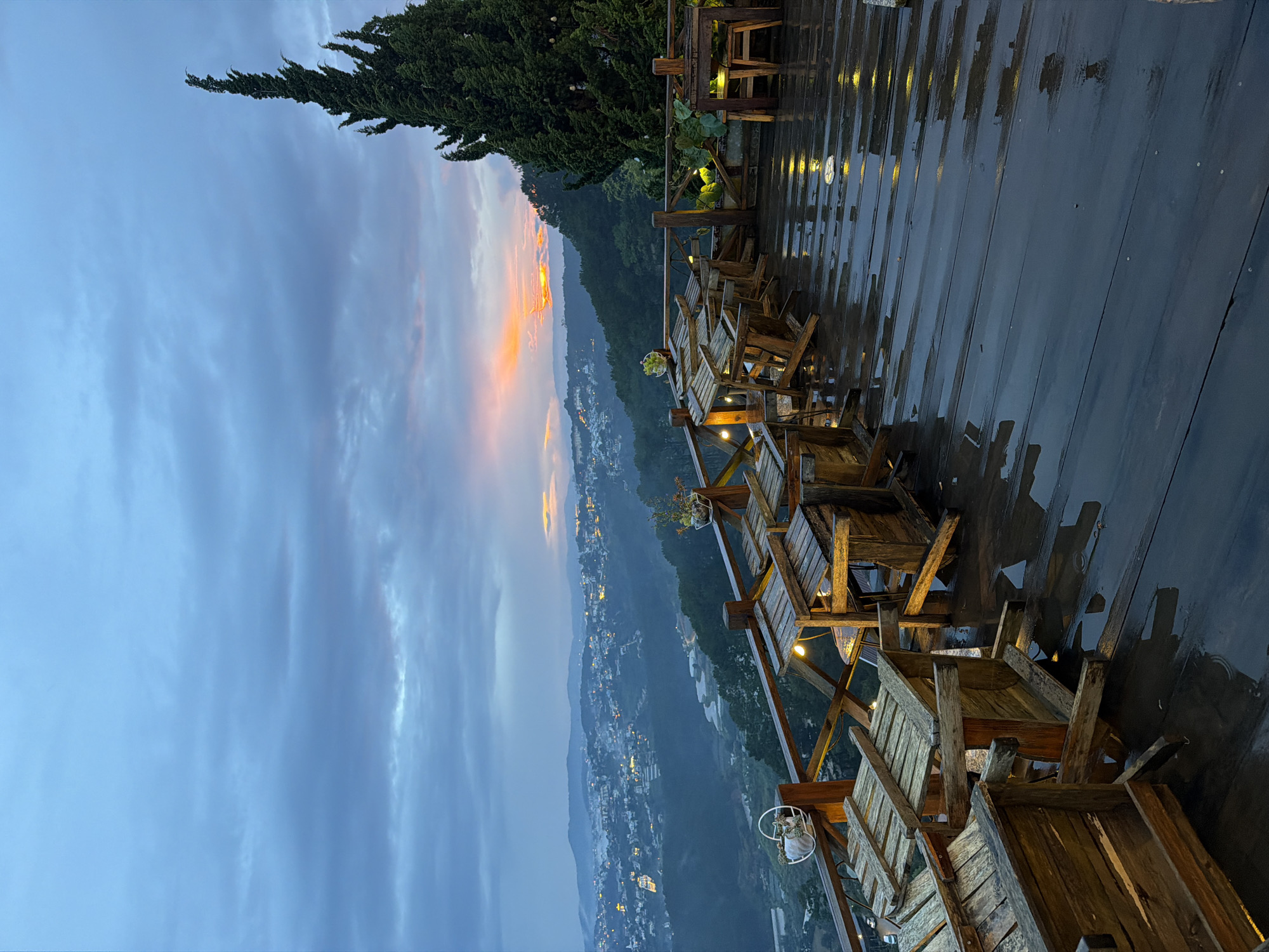 The image depicts an outdoor seating area of a cafe or restaurant overlooking a misty city and rolling hills. The scene is set at dusk or dawn, as evidenced by the soft, diffused light and the glow of city lights in the distance. The sky is a mix of blues and grays with hints of orange and yellow on the horizon, suggesting the sun is either rising or setting.
The foreground features a wooden deck, which appears wet, reflecting the lights of the cafe and the sky. Several wooden tables and chairs are arranged on the deck. Some chairs are stacked, and others are positioned as if ready for use. The furniture has a rustic, weathered look.
In the mid-ground, a wooden railing separates the seating area from a slope covered in trees. Beyond the trees, the city of Xuan Truong - Da Lat, Vietnam, unfolds. The city is nestled among hills, and a light fog or mist hangs over it, softening the details of the buildings and streetlights, which twinkle like scattered jewels.
A tall, conical evergreen tree stands prominently on the right side of the deck, its dark green foliage contrasting with the lighter tones of the sky and the city.
The overall atmosphere is tranquil and somewhat moody, with the wet deck and dim lighting contributing to a serene, almost melancholic feel. No people are visible in the image.