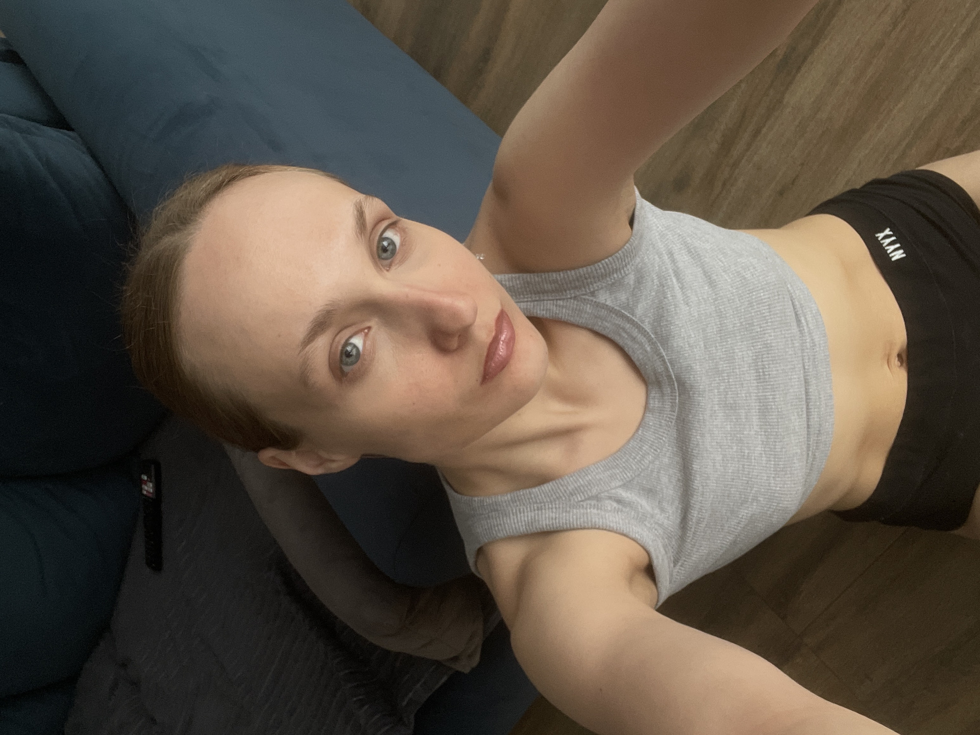 A woman lies on a blue couch and wood floor in what appears to be an indoor setting. She wears a gray tank top and black shorts with the word "YAAN" printed on them. Her hair is pulled back, and she looks toward the camera. A remote control is also on the couch. There is no explicit information to confirm the location of the scene as being in Подшивалово, Russia.