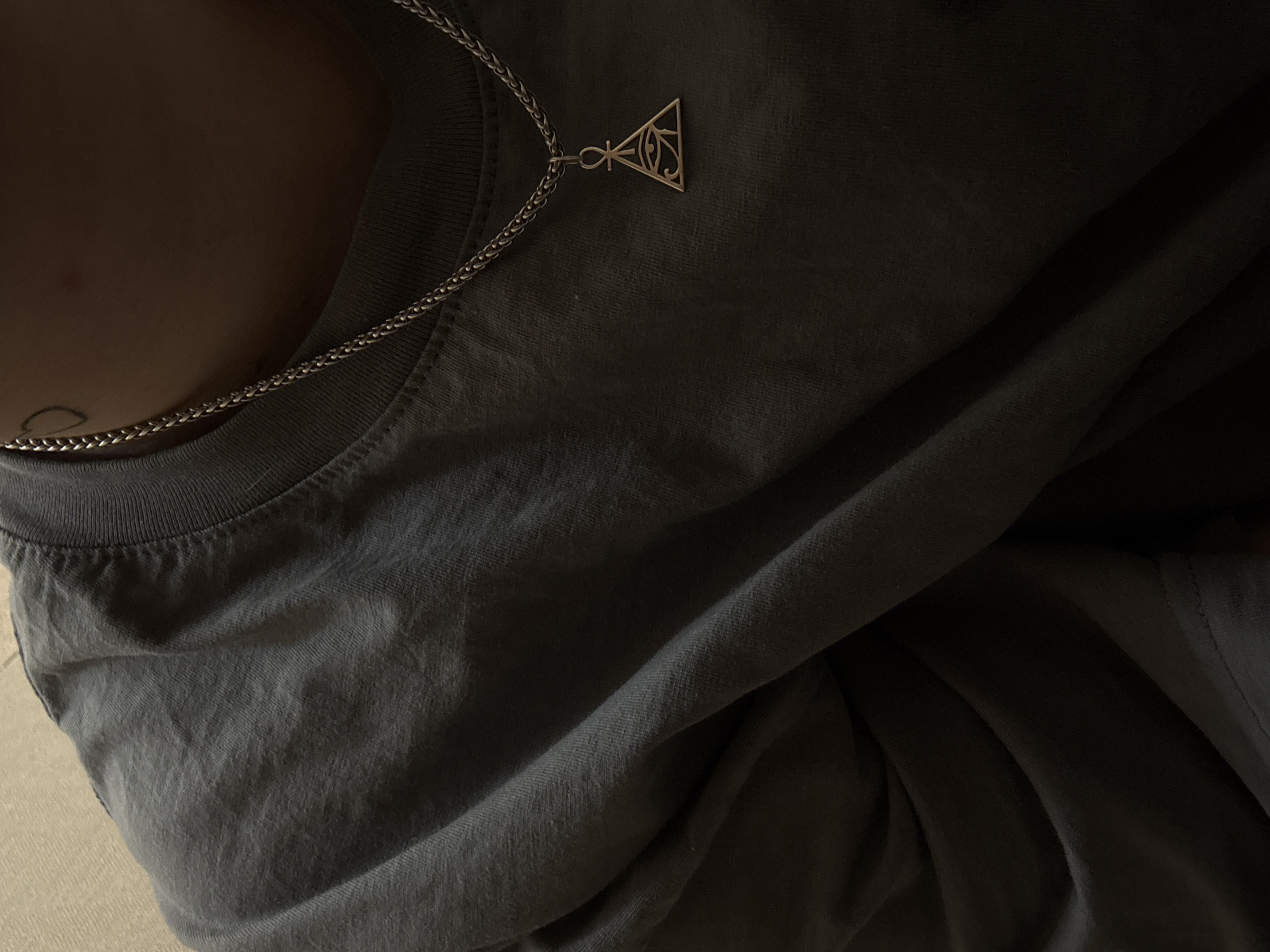 The image shows a close-up of a person wearing a dark grey t-shirt and a silver necklace with a triangular pendant featuring an eye symbol. The person's skin is visible on their neck and shoulder, with a small, faint tattoo of an outline of a heart shape visible on the left side of their neck. The lighting is dim, creating shadows that emphasize the texture of the t-shirt and the detail of the necklace. There are no identifiable elements in the image that would allow for determining its location in Saint Petersburg, Russia, or any specific city context.