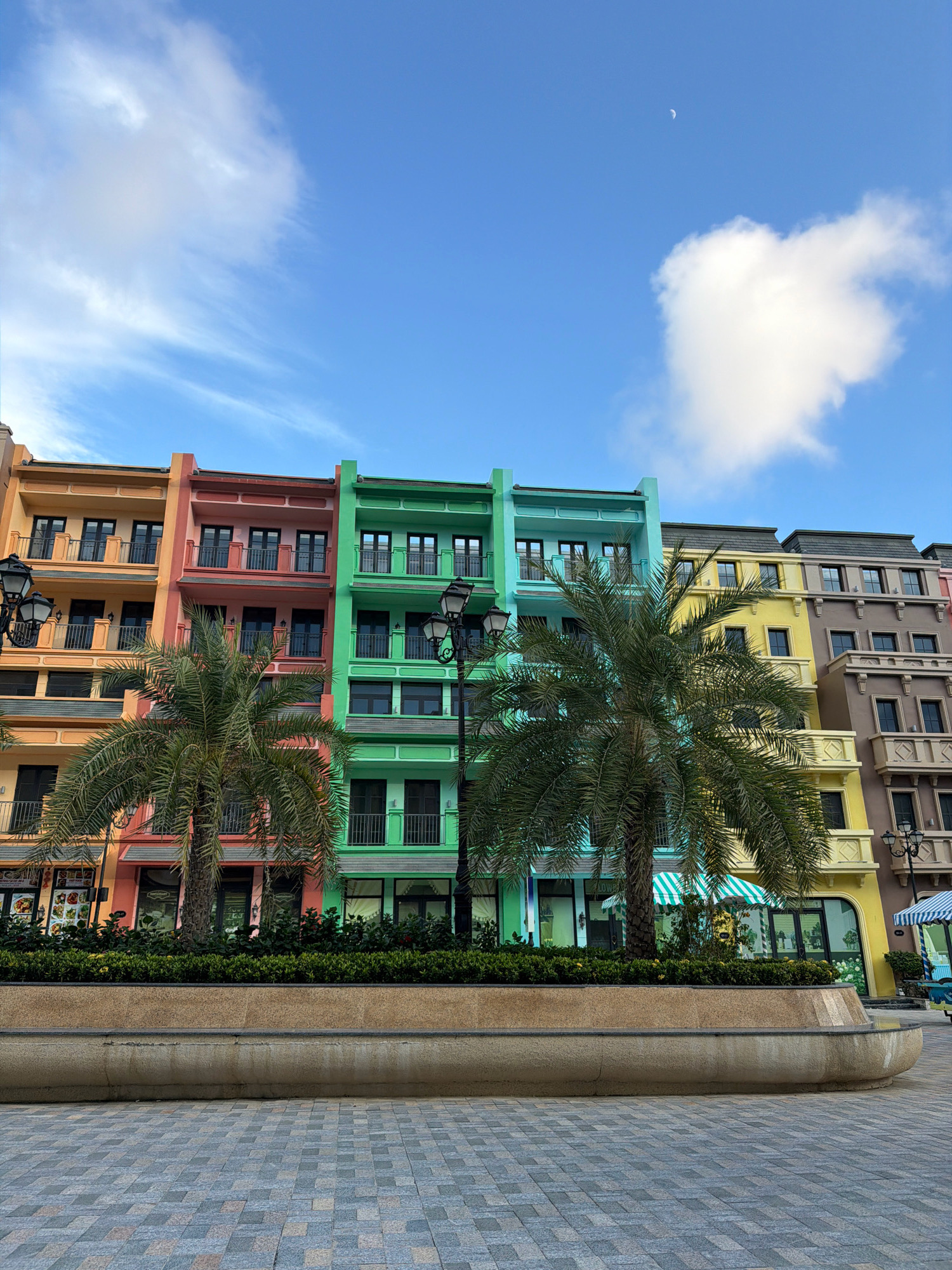 The image captures a vibrant street scene in Nha Trang, Vietnam, showcasing a row of colorful, multi-story buildings. The architecture features brightly painted facades in shades of orange, pink, green, yellow, and beige, with multiple windows and balconies on each level. Palm trees are strategically placed in front of the buildings, adding a tropical element to the urban landscape. The street in the foreground is paved with patterned tiles, and a low, curved concrete planter box runs along the edge of the sidewalk, filled with lush green bushes. In the sky, a crescent moon is visible against a backdrop of blue sky with scattered white clouds. The overall impression is a cheerful and well-maintained street, likely a commercial or residential area designed with an aesthetic appeal.