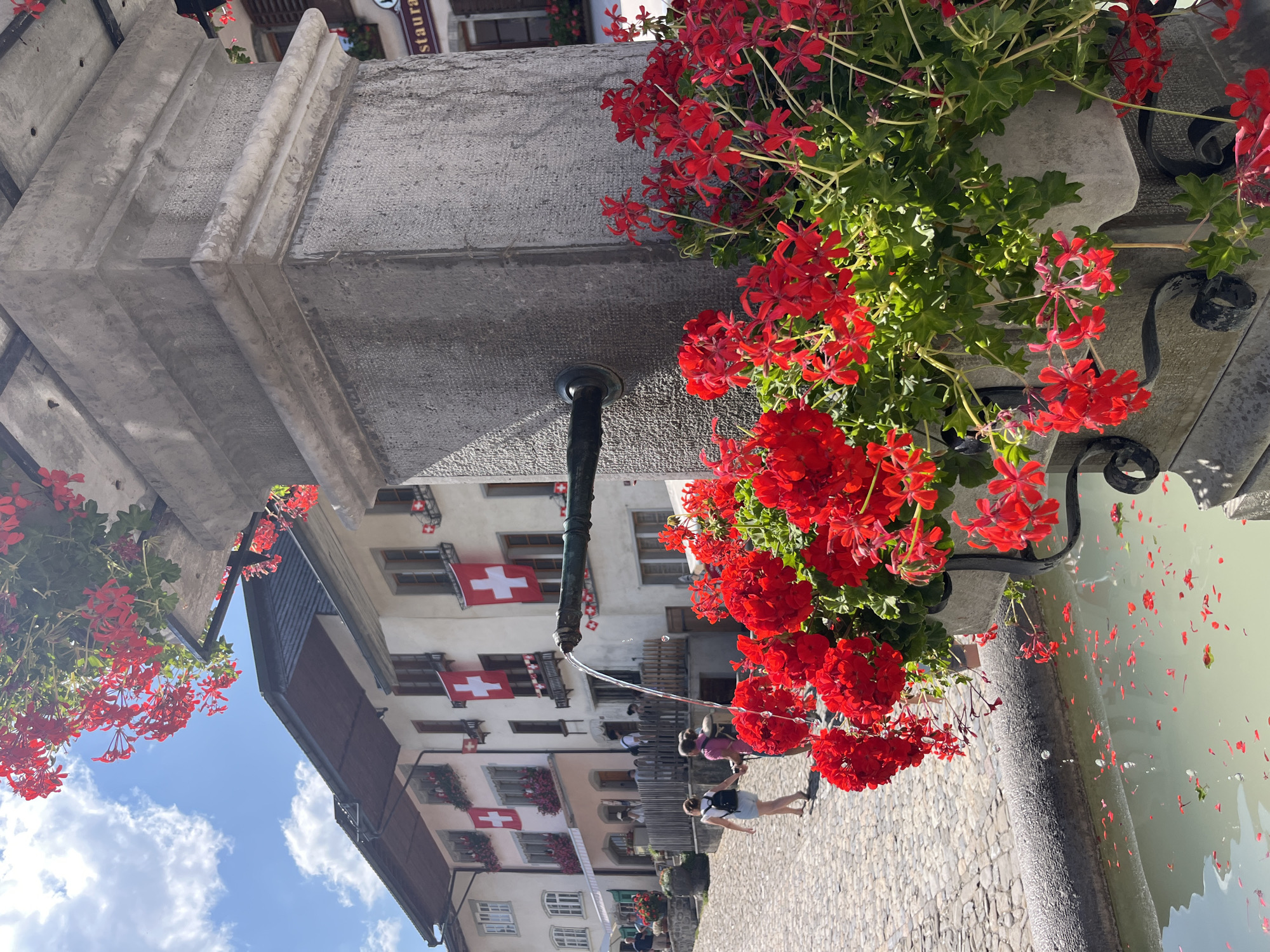 In the city of Epagny, Switzerland, on a cobblestone street, a stone fountain with a black spigot stands beside a building adorned with Swiss flags and window boxes filled with vibrant red flowers. Water flows from the spigot into the fountain's basin, where red flower petals have fallen. The building in the background features traditional architecture with multi-paned windows and awnings. Several people are visible walking on the street, some carrying bags, and a few are standing near the fountain. The sky above is blue with scattered white clouds, and more red flowers are blooming from containers on the fountain and atop the building's stone structure.
