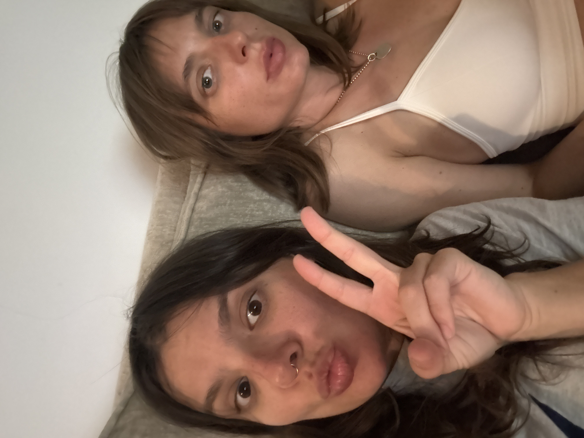 This image captures two adult females indoors, likely a selfie.
The woman on the left, with dark brown hair, dark eyes, and a gold-tone hoop nose ring in her left nostril, is seen with pursed lips in a "duck face" expression. Her right hand is raised, forming a peace sign (V-sign) with extended index and middle fingers. She wears a light-colored garment, possibly a t-shirt or sweatshirt, with a partial dark blue marking visible on her left side.
The woman on the right, with medium brown hair and bangs, and light-colored eyes, has a neutral expression with slightly parted lips, looking directly at the camera. She is wearing a white spaghetti-strap top and a thin, gold-tone chain necklace with a small heart-shaped pendant.
Both women appear to be in their 20s or 30s and are positioned closely together. The background consists of a plain, light-colored upholstered surface, suggesting an indoor setting such as a couch or bed. The lighting is soft and even. No specific visual elements within the image identify the location, however, it is stated to be in the city of Tel-Aviv, Israel.