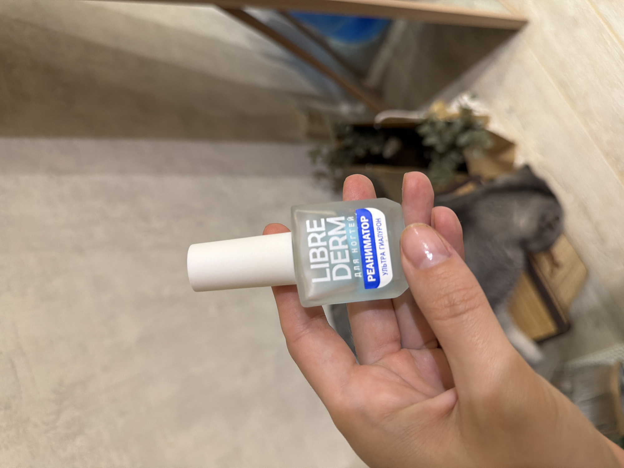 A hand is holding a small bottle of "LIBREDERM" nail reanimator with ultra-hyaluronic acid. The bottle is clear with a white cap and blue and white labeling. The background is out of focus but appears to be an indoor setting with textured walls and some greenery. There is no indication of the city of Semiluki, Russia within the image.