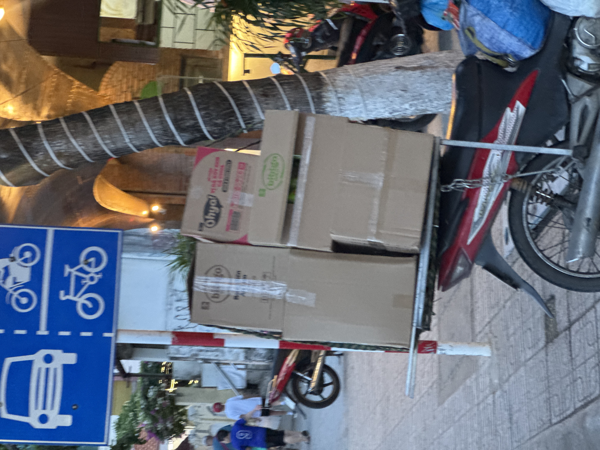 The image shows a street scene in Bac Nha Trang Ward, Vietnam. In the foreground, there is a motorcycle parked on a tiled sidewalk, carrying several cardboard boxes on its rear rack. The motorcycle is red and black, with a blue bag strapped to the back. To the left of the motorcycle, a traffic sign is visible, indicating lanes for cars and bicycles. In the background, there are palm trees and buildings, with some people walking on the sidewalk. The overall scene depicts a typical urban environment in Vietnam, likely during the daytime.