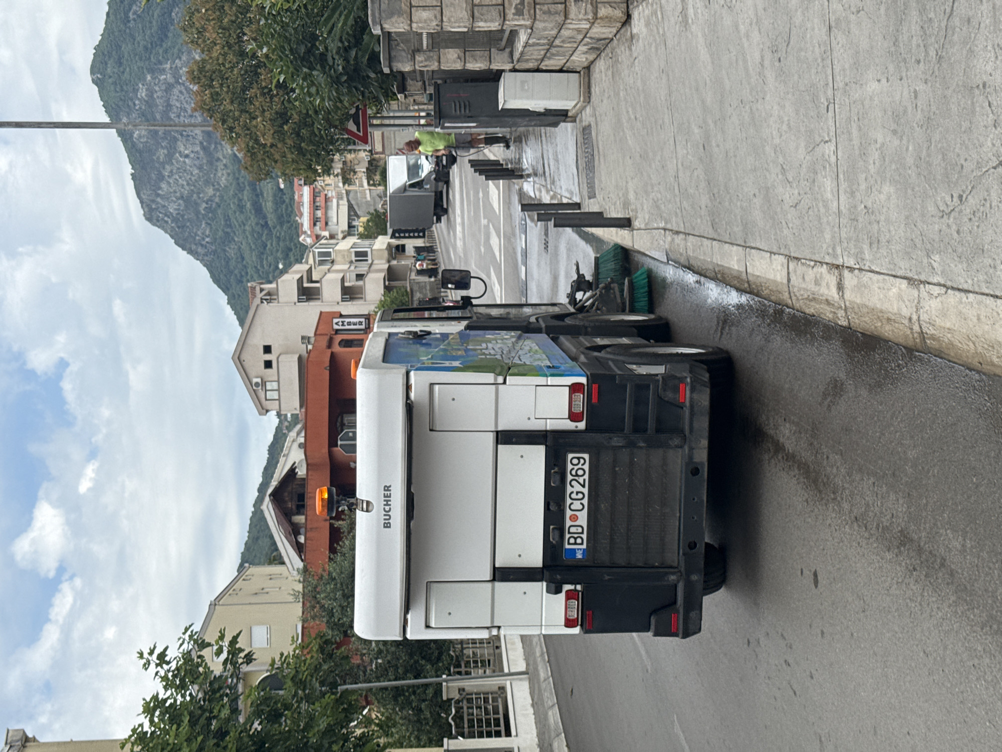 The image displays a daytime street scene in Budva, Montenegro, featuring ongoing cleaning operations. In the foreground, a white and dark grey Bucher street sweeper truck is visible from the rear, parked or moving slowly on a wet asphalt road. The truck's license plate, "BD•CG269," explicitly identifies its origin in Budva, Montenegro, with "MNE" also visible. An orange warning light is illuminated on the truck's front left side.
Adjacent to the truck, a concrete sidewalk is also wet, extending towards a person in a bright green shirt who is actively using a pressure washer. This individual is seen spraying water onto the sidewalk, confirming the street cleaning activity. Black bollards are spaced along the sidewalk, defining the pedestrian area.
The background reveals a dramatic natural setting dominated by a massive, steep mountain covered in dense green vegetation. Below the mountain, several multi-story buildings line the street, with diverse architectural styles. One building has a sign with the word "AMBER" clearly legible. The sky above is partly cloudy with patches of blue. The overall atmosphere suggests an active, well-maintained urban environment set against a striking natural landscape.