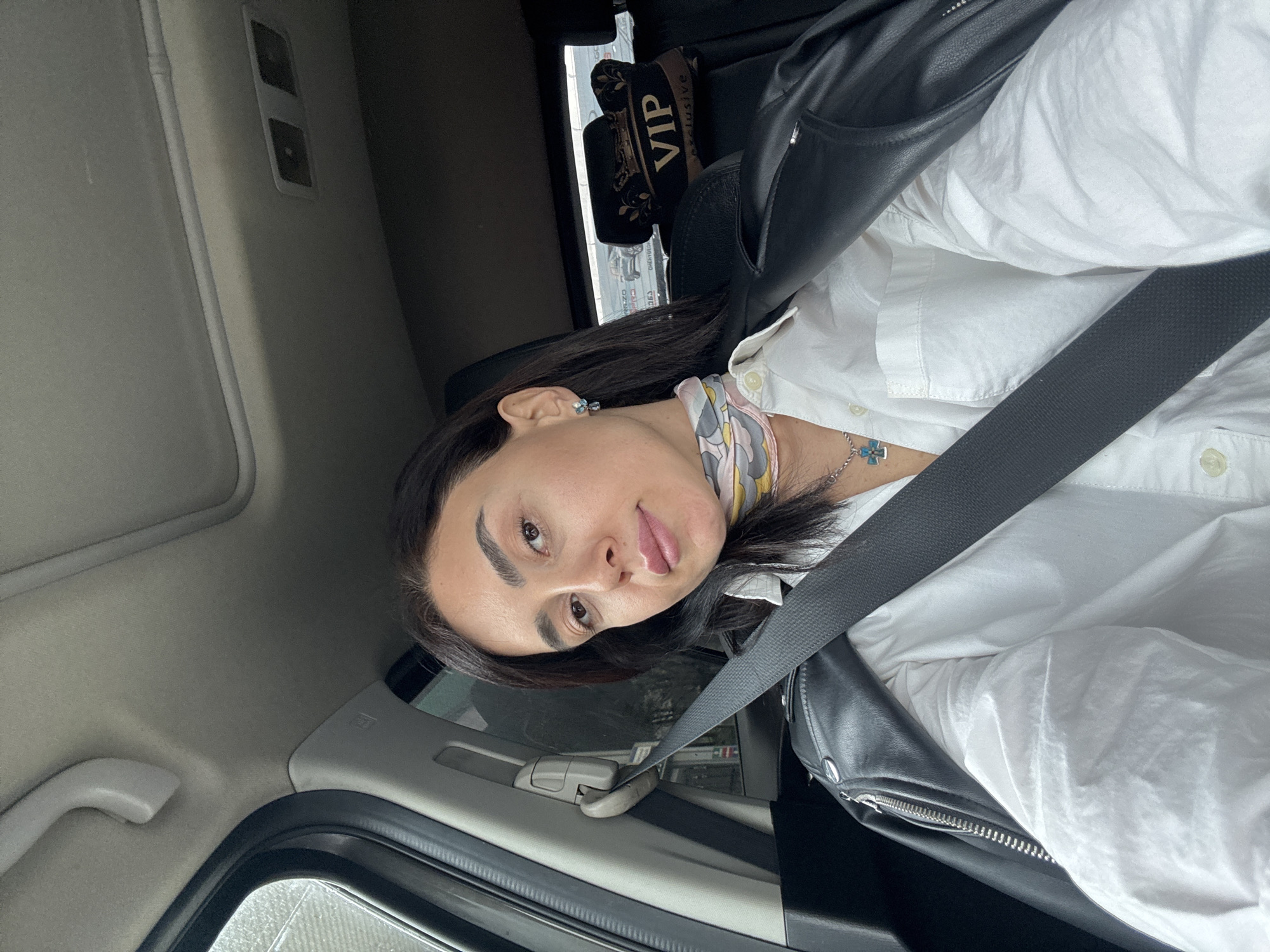 A woman with dark hair is seated inside a vehicle, wearing a white button-down shirt, a black leather jacket, a patterned scarf, and a seatbelt. She has earrings and a necklace. A "VIP Exclusive" sign is visible in the background. The scene appears to be within a car, possibly in Tbilisi, Georgia, based on background signage visible through the window. The angle of the photograph is from the front and to the side, slightly angled up.