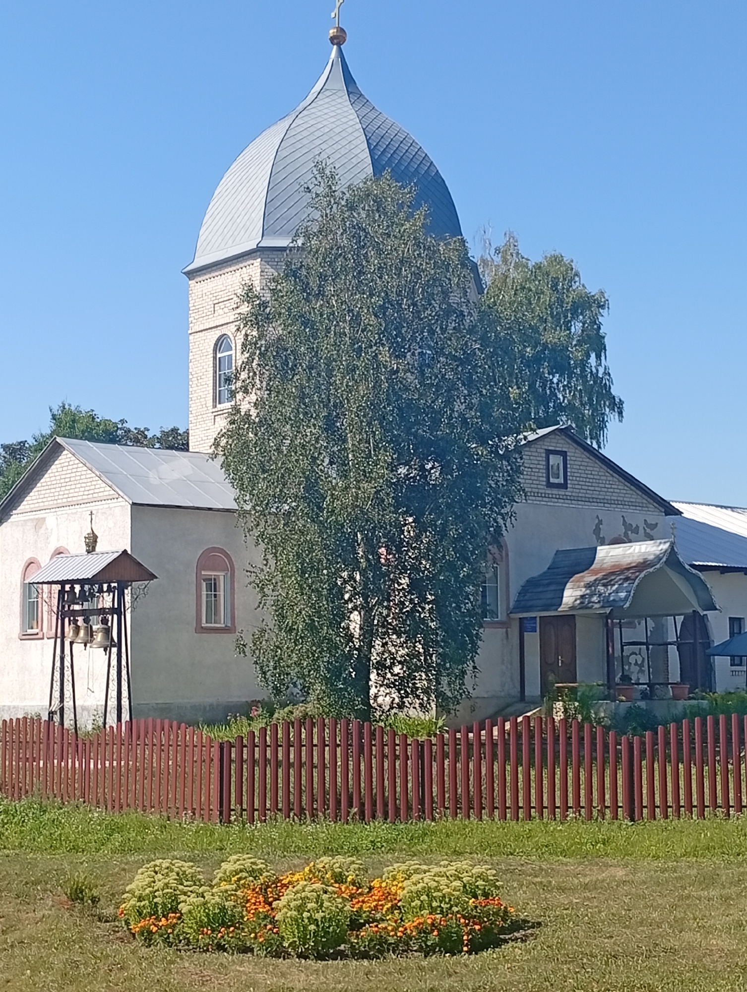 The image shows a church in the village of Khomutets, Ukraine. The church has a light-colored brick tower with a grey dome topped with a cross. The main body of the church is also light-colored and has a light-colored metal roof. In front of the church is a red wooden fence. To the left of the church is a bell tower with several bells. In the foreground, there is a flower bed with orange and yellow flowers. The sky is clear and blue.