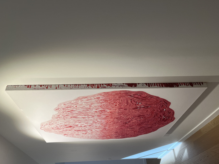 The scene captures a large, vertically-oriented abstract painting mounted on a plain white wall within an indoor corridor or gallery space in Paris, France. The artwork prominently displays an irregular, elongated form composed of dense, vertical, wavy, and dripping lines in varying shades of red and pink against a white background. The visible right-hand edge of the canvas exhibits red paint drips extending down its white side. An overhead light source illuminates the upper section of the artwork, casting a soft glow. A distinct diagonal beam of natural or artificial light strikes a distant portion of the left wall and a light-toned wooden floor. No individuals are visible, and no specific actions or interactions are occurring. The environment presents as a clean, modern interior.