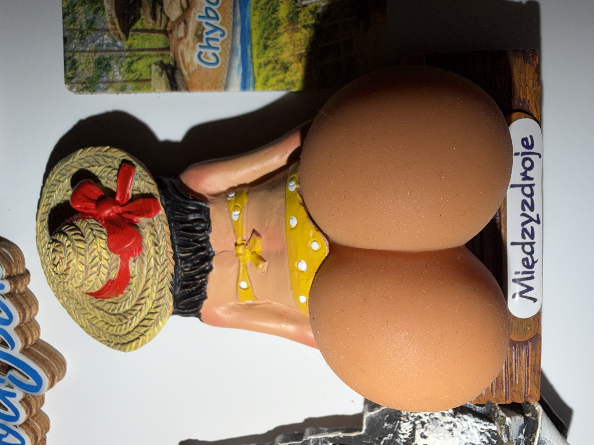 This media file depicts a novelty item, likely a magnet or figurine, shaped like a woman wearing a straw hat adorned with a red ribbon. She is dressed in a yellow polka-dot outfit. The figurine has two large, round, light brown objects that resemble eggs attached to her. The background shows a postcard-like image of a rocky, natural landscape with trees.
Visible text includes "Chyba" on the postcard image and "Międzyzdroje" written on a white label attached to a wooden backing behind the figurine. "Międzyzdroje" is a resort town in northwestern Poland. The overall presentation suggests a souvenir related to a Polish location. The lighting in the image is bright, casting a distinct shadow from the figurine, indicating it was taken in a well-lit environment, possibly indoors. There are no clear indicators of the time of day or weather conditions. The scene is static and does not depict an activity in progress.