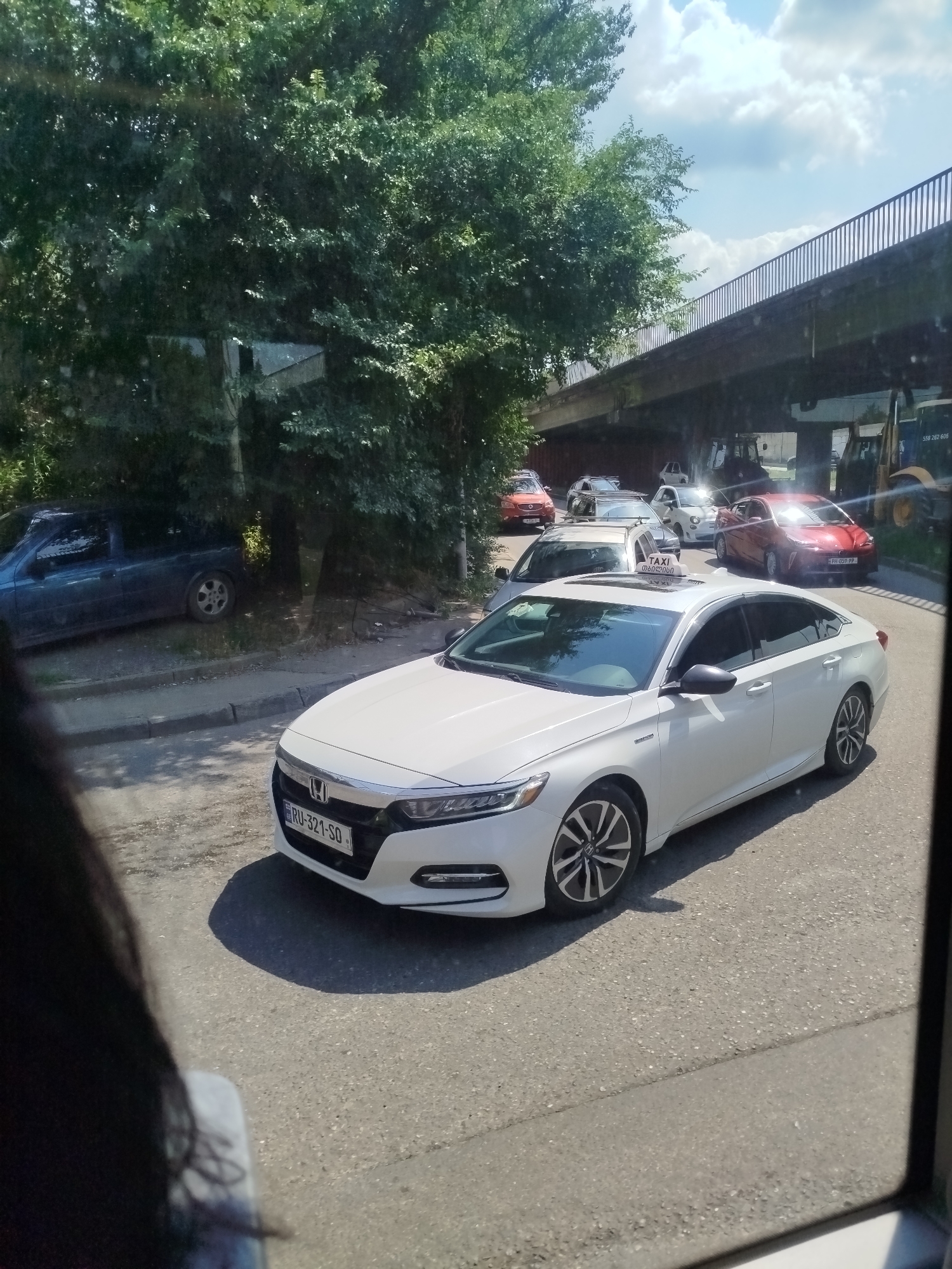 The image shows a white car, possibly a taxi, with a "TAXI" sign on its roof, driving on a road. The car is in the foreground, and other cars are visible behind it, suggesting a traffic situation. The background features lush green trees and a concrete overpass with a railing. The sky is partly cloudy, indicating a bright day. The scene appears to be an urban or suburban environment during daylight hours. The license plate of the white car is visible and reads "RU-321-SO". The image is taken from inside another vehicle, as suggested by the window frame on the right side and the partial view of a person's dark hair in the bottom left corner. The overall mood is neutral, capturing a typical street scene.