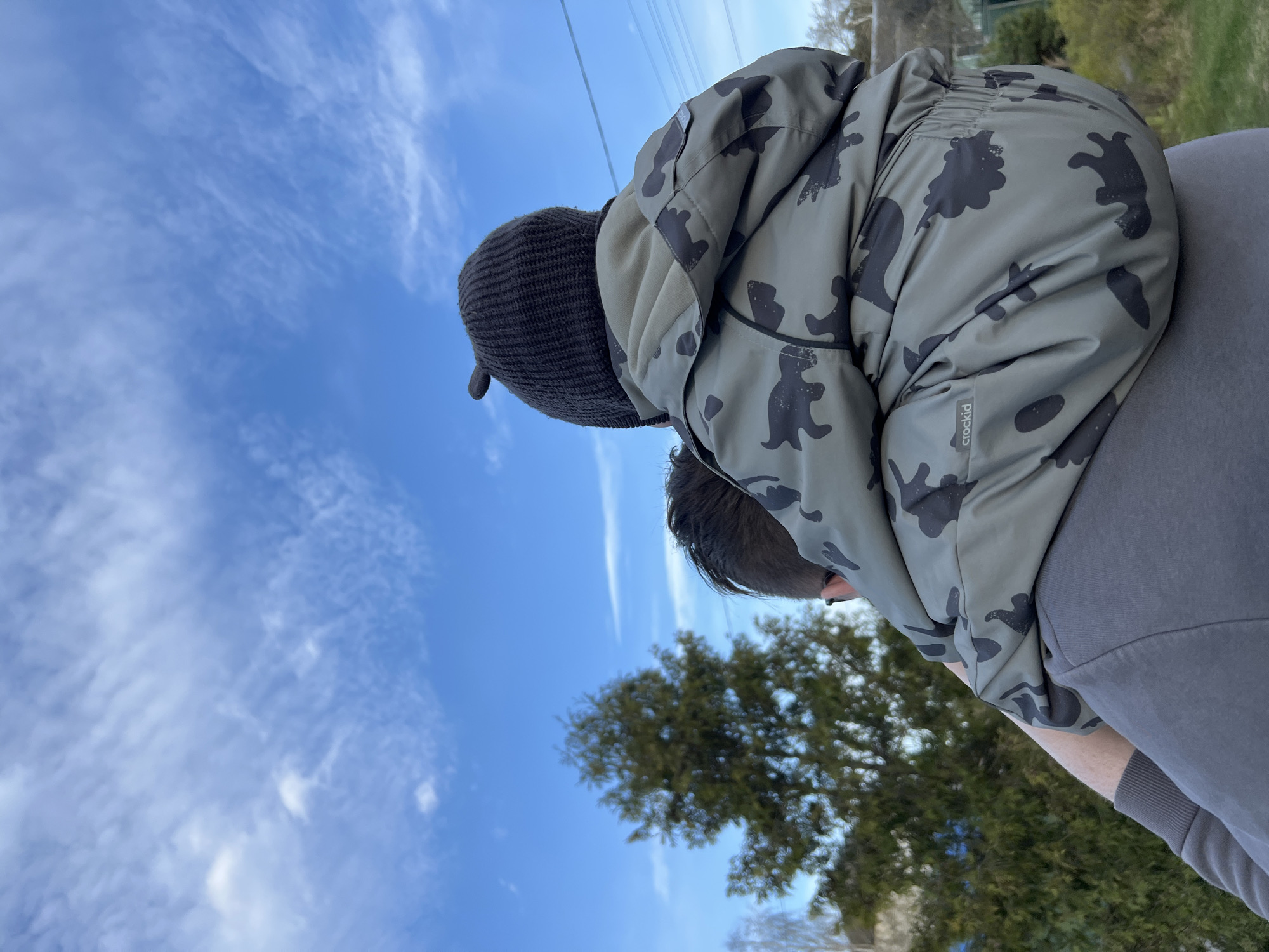 A person is giving a piggyback ride to a child outdoors. The child wears a dark beanie and a patterned jacket with dinosaur motifs. The adult wears a gray shirt. The background features a blue sky with scattered clouds and trees. The photo appears to have been taken from a low angle. The location is Razdolye, Russia.