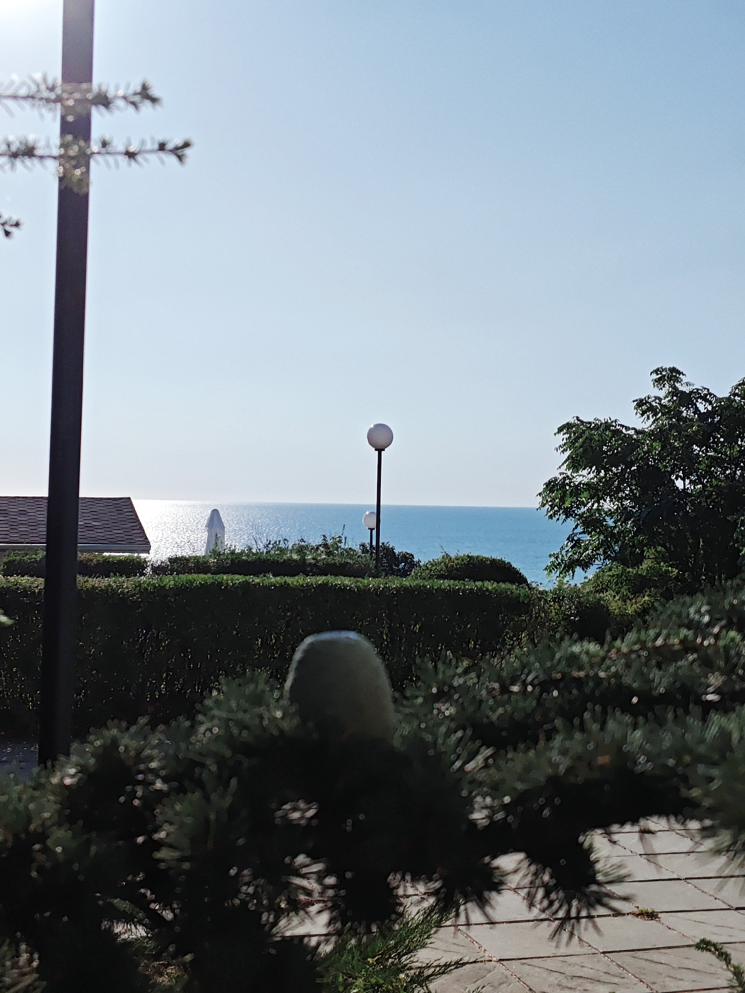 In an outdoor scene, a black lamppost stands to the left, with blurred evergreen branches in the foreground. A tiled walkway is visible behind the branches. Further back, a low-trimmed hedge forms a line across the scene. Beyond the hedge, a building with a brown shingled roof is visible on the left. In the middle ground, a tall, white, umbrella-like object stands on the edge of a body of water, which reflects the bright sunlight. Two lampposts with spherical tops are positioned behind the hedge, with the sea stretching to the horizon under a clear blue sky. A large, dark green tree occupies the right side of the image. The overall scene appears to be a coastal location in a bright, sunny environment.