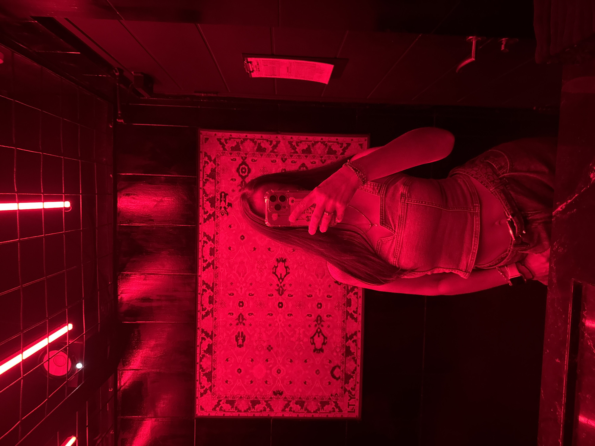 The image shows a woman taking a selfie in a bathroom with red lighting. She is standing on a patterned rug and holding a phone in her right hand. The bathroom has black tiled walls on the left, a wooden wall behind the rug, and black fixtures on the ceiling, including a light and a hook. A bright red neon light is visible on the left wall. The location is a bathroom in Kuta Selatan, Indonesia.