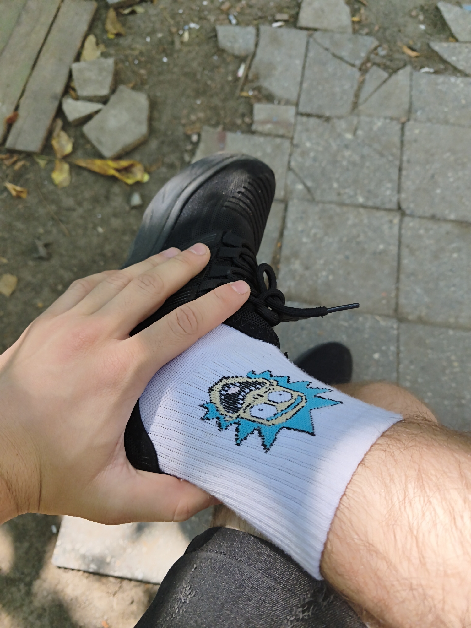 A person's hand, with fair skin and visible veins, is shown adjusting a white sock with a cartoon character print, possibly Rick Sanchez from the show Rick and Morty, on their ankle. The sock is worn with a black sneaker. The background suggests an outdoor urban setting, with cracked concrete paving stones and some fallen leaves, indicative of an area that may be undergoing repair or is somewhat neglected. The scene is lit by natural daylight. The specific location in Chișinău, Moldova cannot be determined from the visual information provided.