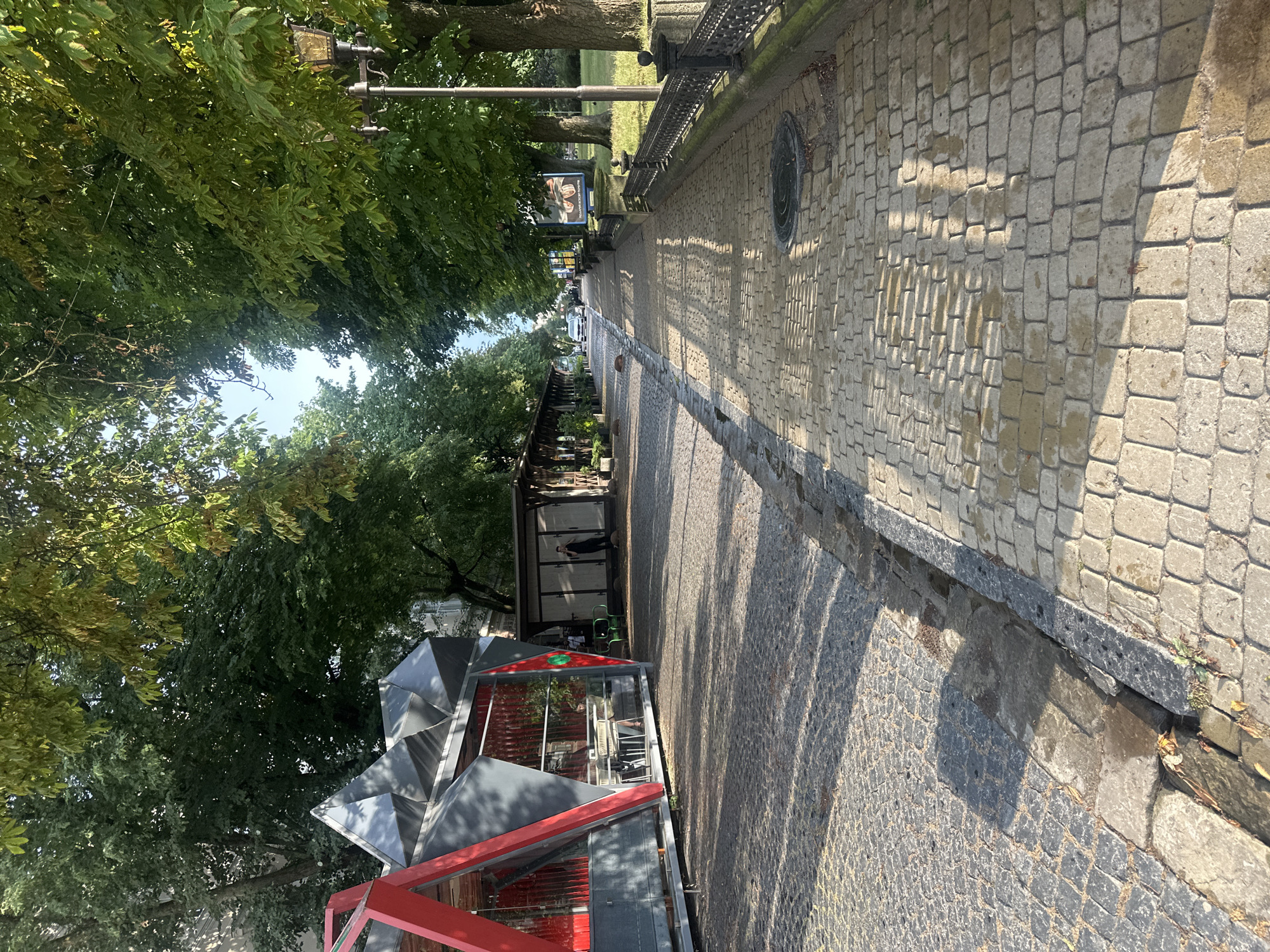 The image shows a street in Ternopil, Ukraine, likely a promenade or a street with pedestrian and vehicle access. On the left, there is a modern-looking structure with red and gray geometric elements, possibly a cafe or a kiosk. Tables and chairs are visible inside through the glass. Adjacent to this structure are several smaller, wooden kiosks with awnings, creating a row of shops or food stalls. A person in dark clothing is standing near one of these stalls.
The ground is paved with cobblestones, with a distinct path made of lighter-colored rectangular stones for pedestrians, and darker, more irregular cobblestones on the side, likely for vehicles or a service area. A drainage ditch runs along the edge of the pedestrian path.
On the right side of the image, a black metal fence with decorative elements separates the walkway from a grassy area. A street lamp with an ornate design stands near the fence. Tall trees with lush green foliage line both sides of the street, casting shadows on the pavement. In the background, a few cars are visible, suggesting that this is a public street. A large poster is mounted on a pole near the fence. The sky is visible between the tree branches and appears to be a clear blue. The lighting suggests it is daytime, with the sun casting distinct shadows.