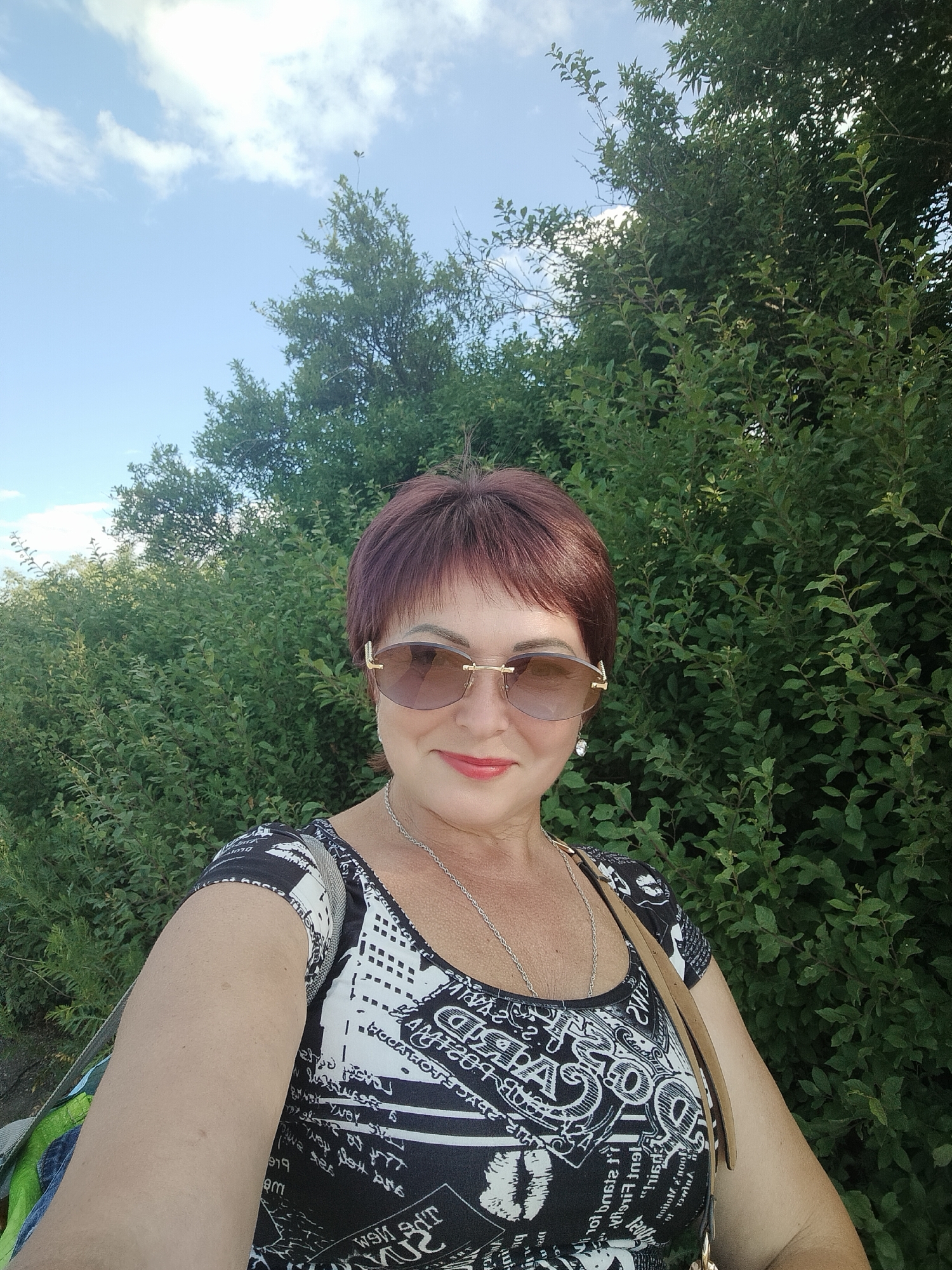 A woman with short, reddish-brown hair and sunglasses takes a selfie outdoors. She is wearing a black and white patterned top and a silver necklace. Her left arm is visible, and a purse strap crosses her chest. The background is filled with lush green foliage, suggesting an outdoor setting with trees and bushes. The sky above is a clear blue with some white clouds, indicating daytime and fair weather. The woman is smiling slightly, conveying a relaxed and pleasant demeanor. There is no visible text in the image. The location context is Lykhove, Ukraine.