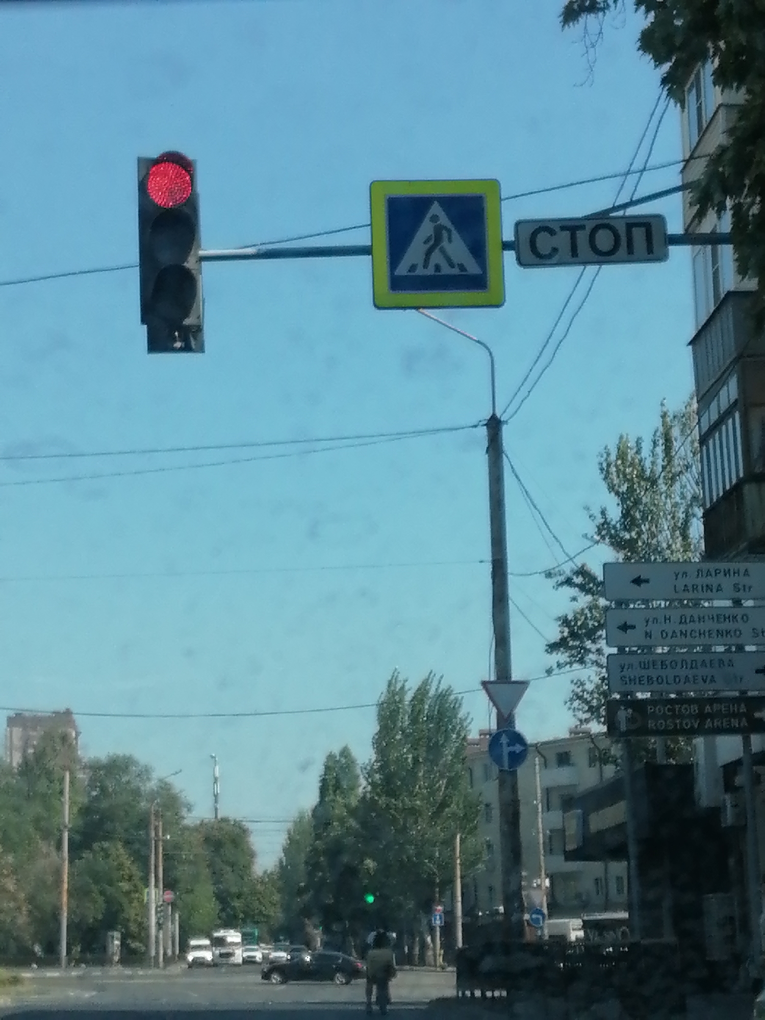 The image depicts a street scene in Rostov-on-Don, Russia, under a clear, bright sky. In the foreground, a traffic light shows a red signal. To the right of the traffic light, a pedestrian crossing sign is visible, featuring a blue square with a white outline of a person walking. Adjacent to the pedestrian crossing sign is a rectangular sign with the word "СТОП" (STOP) in black capital letters on a white background, indicating a stop sign. Further to the right, a series of directional signs are mounted on a pole. These signs indicate directions to various streets, including "ул. ЛАРИНА LARINA Str", "ул. Н. ДАНЧЕНКО N. DANCHENKO Str", "ул. ШЕБОЛДАЕВА SHEBOLD AEVA Str", and "РОСТОВ АРЕНА ROSTOV ARENA".
In the mid-ground, several vehicles are present on the road, including a white truck and several cars. Trees line the street, and buildings can be seen in the background. The presence of multiple traffic signs and the urban setting suggest this is a typical intersection within the city.
In the lower right corner, a person is partially visible, walking away from the camera. The overall impression is a snapshot of urban traffic flow and street signage.