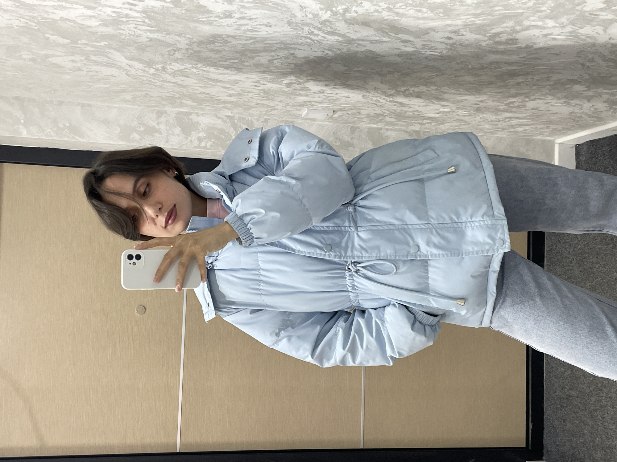 A woman is taking a selfie in a room. She is holding a white smartphone. She is wearing a light blue puffer jacket and gray pants. The room has beige walls and a gray and white patterned ceiling. A black-framed mirror is visible. It is not possible to determine the exact location within Izhevsk, Russia, without additional information.
