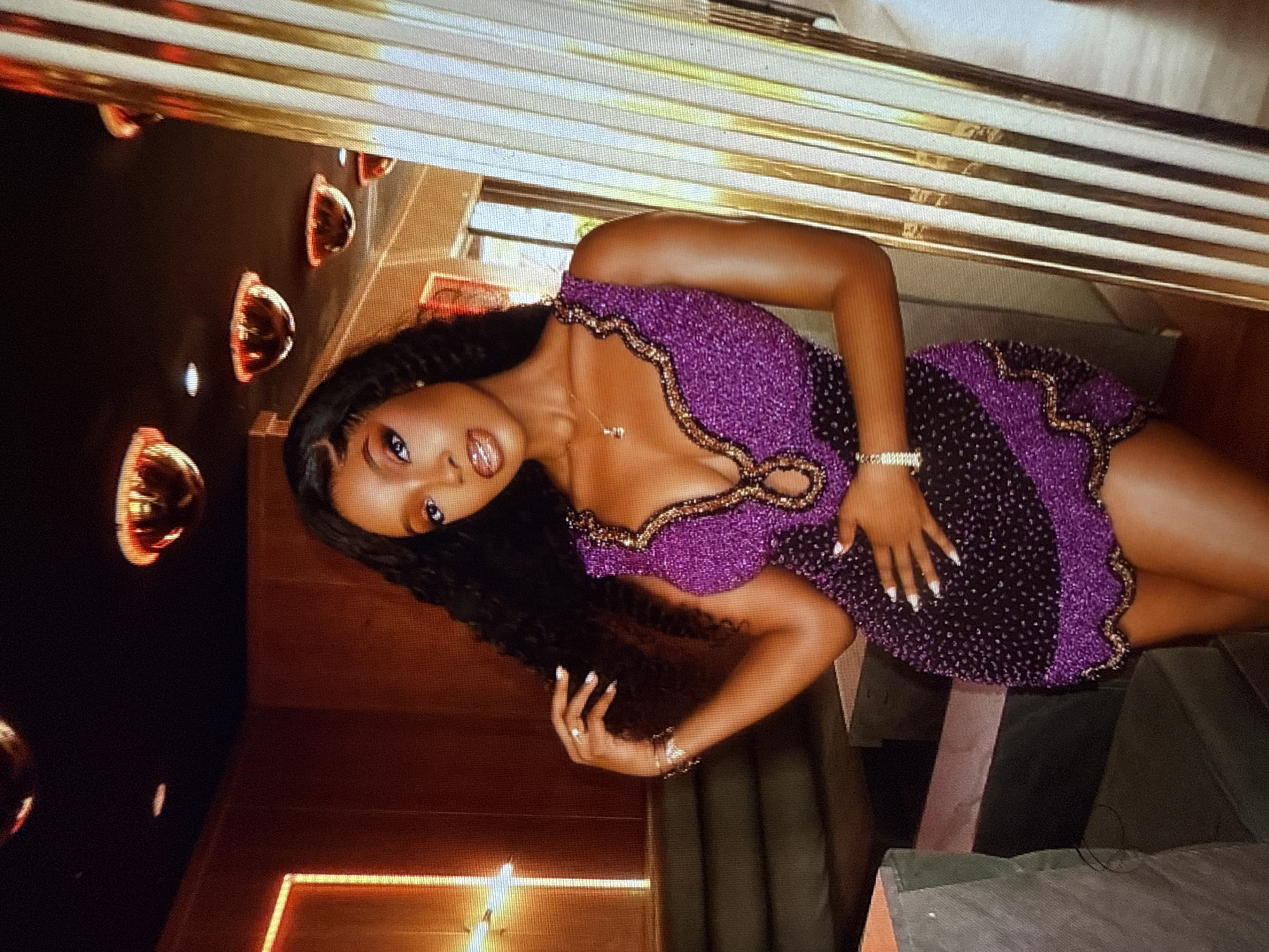 A Black woman with long, dark, curly hair stands in what appears to be a dimly lit, upscale establishment. She is wearing a sleeveless, short, fitted dress adorned with purple and black beads, and gold trim around the neckline and hem. The dress features a deep V-neckline with a cut-out detail. She has on bracelets on her left wrist and a delicate necklace. Her left hand is posed near her hair, and her right hand rests on her stomach. Her makeup is noticeable, with bold eyeshadow and lip color. In the background, there are dark wood-paneled walls, plush seating, and decorative ceiling lights that resemble upside-down bowls with red interiors. To her right, a fluted column with gold and white stripes is visible. The overall ambiance suggests a night out in a stylish venue. While the image is captured in Abuja, Nigeria, this specific location within the city is not identifiable from the visual information alone.