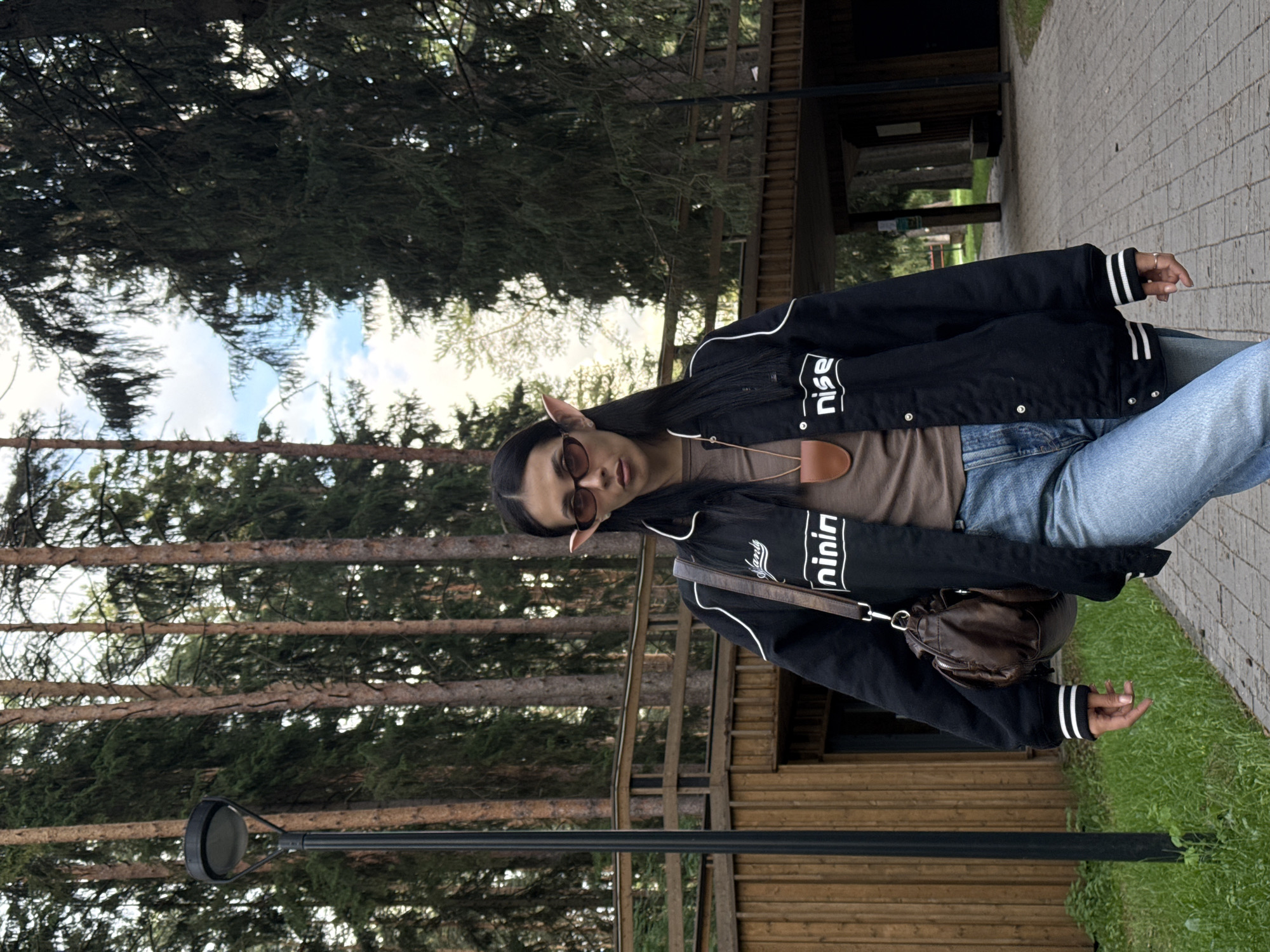 A full-length shot depicts a woman standing outdoors, likely in a park or wooded area. She wears a black bomber jacket with white trim, blue jeans, and a brown top. A brown leather bag hangs over her shoulder. She has long dark hair, sunglasses, and pointed elf-like ears. Behind her are tall trees and a wooden structure. The setting appears to be a park with a paved walkway. Though the precise location in the city of Votkinsk is unspecified.