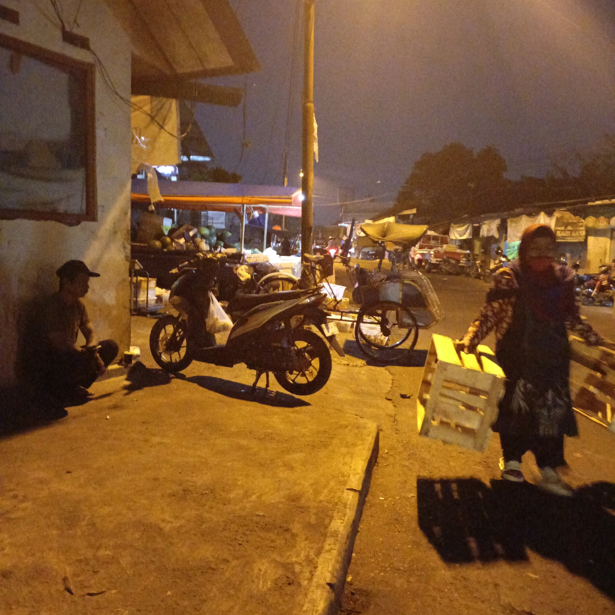 The image depicts a nighttime scene at a roadside market or street food stall in Salatiga, Indonesia. A man wearing a cap and dark clothing is seated on the ground near a building, appearing to rest. In the background, a motorcycle is parked, and a tricycle cart is visible. A woman, wearing a headscarf and a patterned top over dark clothing, is walking towards the right of the frame, carrying a wooden crate. The environment is illuminated by artificial lighting, casting long shadows and giving the scene a warm, yellow-orange hue. Various market stalls and items, such as fruits and bags, are present in the background, suggesting commercial activity. The ground is paved, and there are some debris and stains visible. The overall impression is that of a typical evening in a bustling Indonesian urban area.