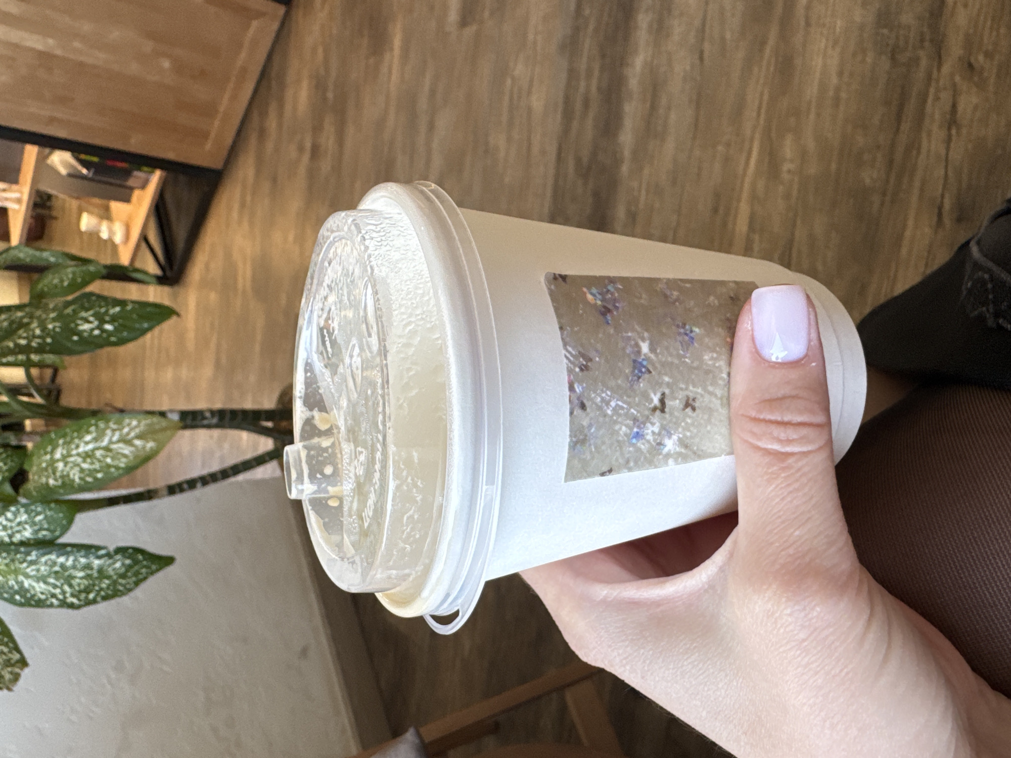 A hand holds a covered disposable cup. The cup is clear plastic with a white paper sleeve. The lid is clear with a sip opening. The person's thumb holds the cup against the white paper sleeve. The thumb has a pale purple manicure. Brown leggings are visible. The background includes a wooden floor and a houseplant. The location is Chaikovsky, Russia.