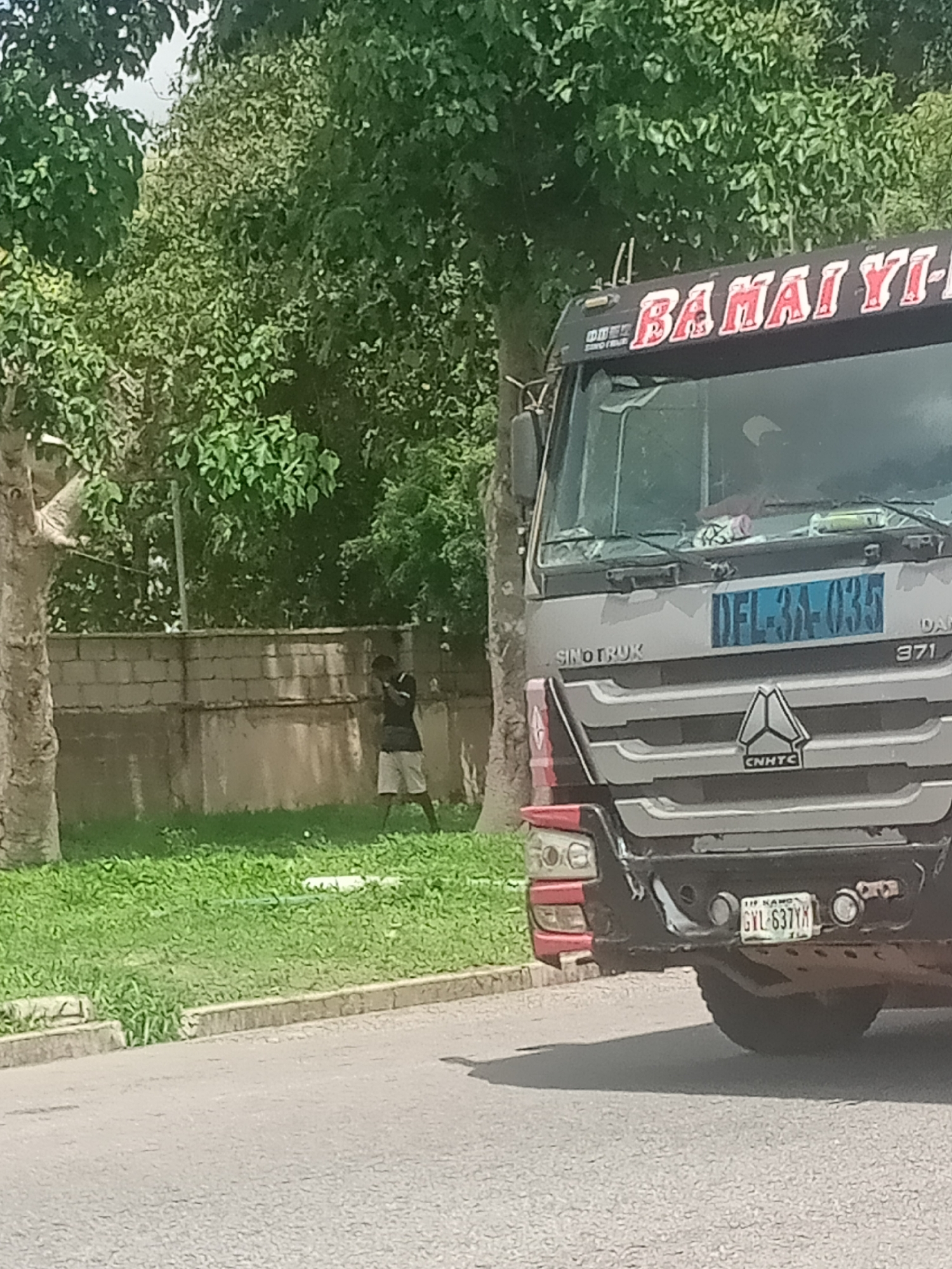The image shows a street scene in Jalingo, Nigeria, with a large truck parked on the right side of the frame. The truck is silver and black, with red lettering on the top that reads "BAMAI YI-". A license plate on the front of the truck is visible and reads "DEL-391-035". The truck has a prominent CNNHTC logo on its grille. In the background, a man wearing a dark shirt and light-colored shorts stands near a low stone wall, holding what appears to be a phone to his ear. He is facing away from the camera. Green grass covers the area between the man and the road. Trees with lush green foliage are visible behind the wall and above the truck. The road in the foreground is paved and gray. The license plate on the rear of the truck, partially visible at the bottom right, reads "GYL-637YX".
