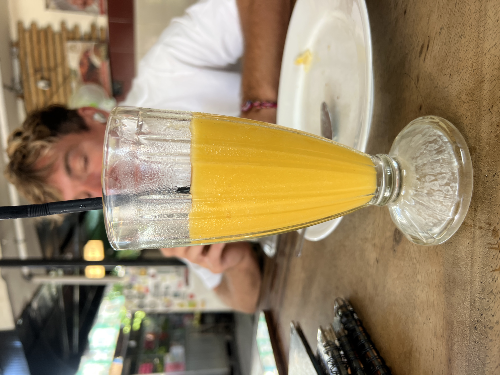 A close-up shot shows a glass filled with orange juice and a black straw. The glass is on a wooden table. In the background, a person wearing a white t-shirt is visible, with their face blurred and eyes closed. The setting appears to be an outdoor cafe in Kuta, Indonesia, possibly The Onion Collective, given the decor. The table also has a white plate with a spoon, and a few notebooks are stacked on the left side of the frame. The overall atmosphere suggests a relaxed, casual dining experience.