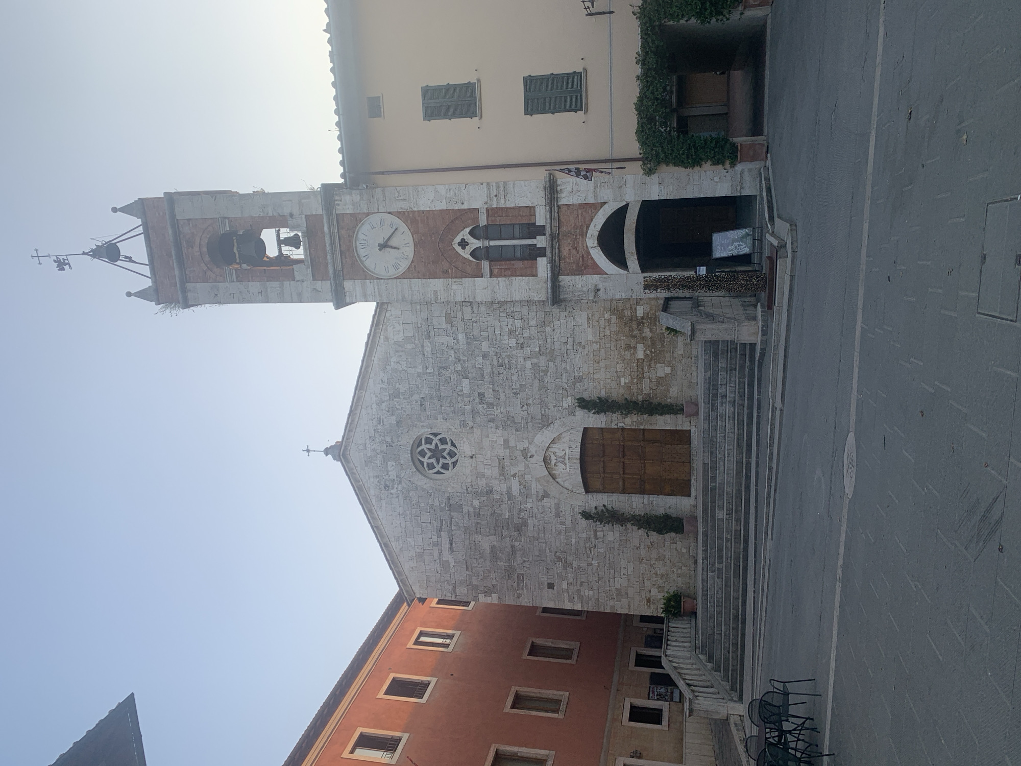 The image shows a view of the Piazza della Libertà in San Quirico d'Orcia, Italy. In the center is the Church of San Quirico, a stone building with a large wooden door and a rose window above it. To the left of the church is a taller building with orange facade and multiple windows. To the right of the church is a bell tower, also made of stone and brick, with a clock face. The sky is clear and light blue. In the foreground of the image, there is a paved square with a few tables and chairs. The overall scene depicts a quiet town square in Italy.