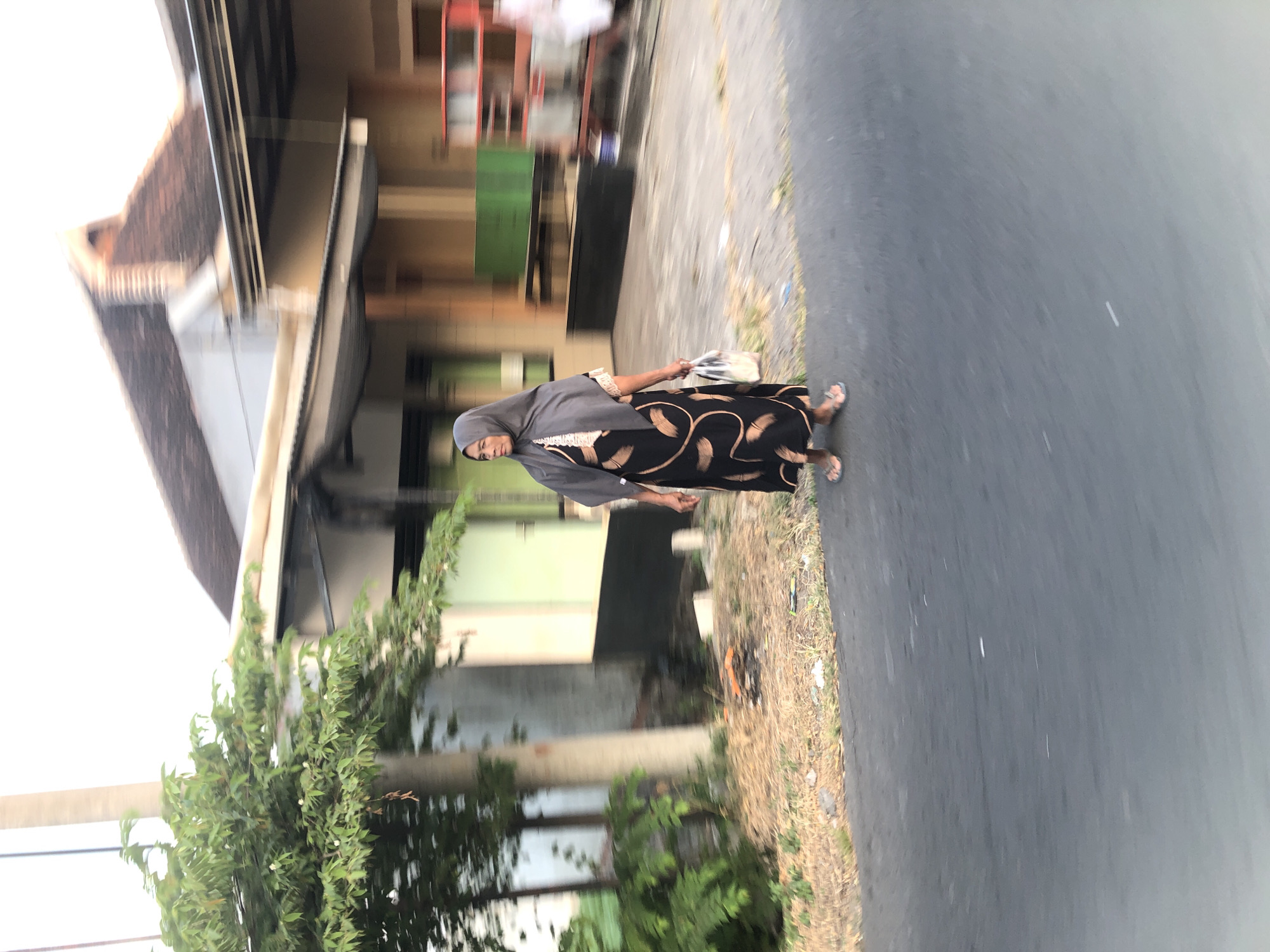 In the city of Glagah Arum, Indonesia, a woman wearing a grey hijab and a black dress with a tan leaf pattern walks on a paved road. She is carrying a translucent plastic bag in her right hand. To her left is a tree with green leaves and a light-colored pole. Behind her, a building with a brown, tiled roof and beige walls is visible. The scene is slightly blurred, suggesting movement.