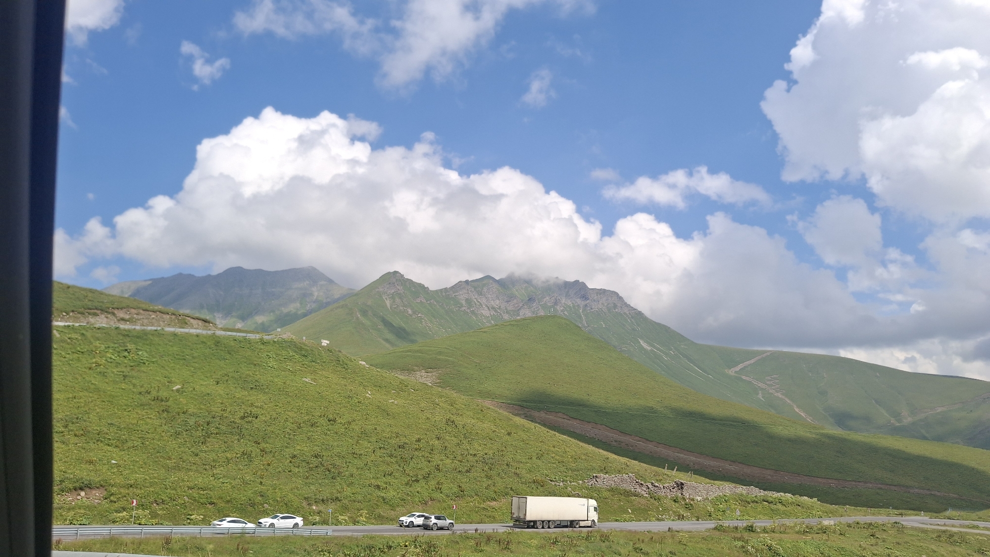This image captures a scenic view of the Georgian Military Highway, near the ski resort town of Gudauri, Georgia. The foreground features a road with several vehicles, including two white cars and a white semi-truck. The road is bordered by a guardrail and lush green fields that slope upwards towards imposing mountains. The mountains in the background are rugged and green, with some areas appearing rocky and others covered in dense vegetation. The sky above is a vibrant blue, dotted with fluffy white clouds, suggesting a pleasant, possibly sunny day. The overall impression is one of a beautiful, natural landscape with human activity present on the highway.