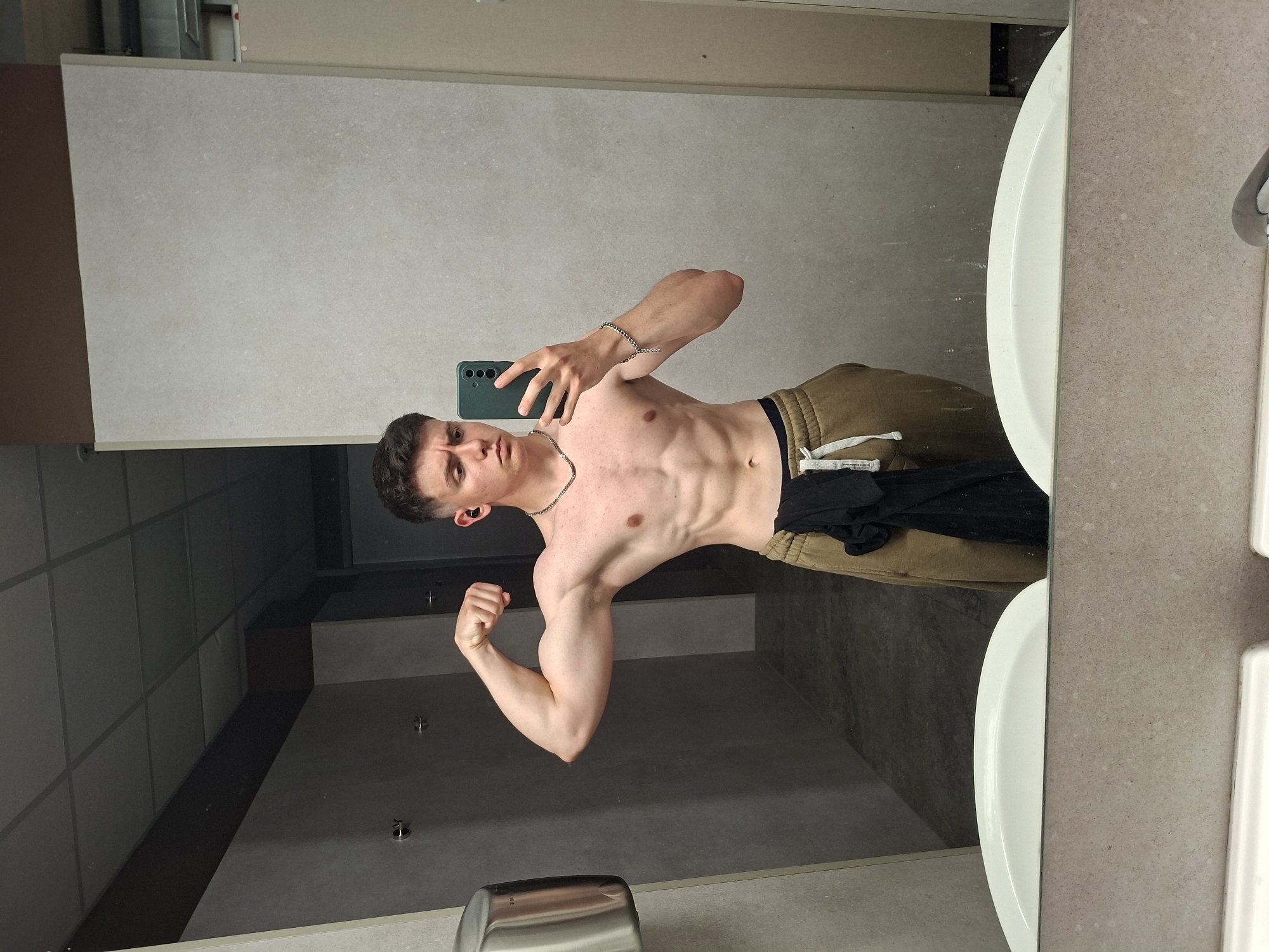 A shirtless young man is taking a selfie in what appears to be a public restroom. He is wearing beige sweatpants and a silver chain. He is flexing his bicep in the mirror. There are white sinks, a gray countertop and a gray wall behind him. The location is not identifiable as being in Plovdiv, Bulgaria.