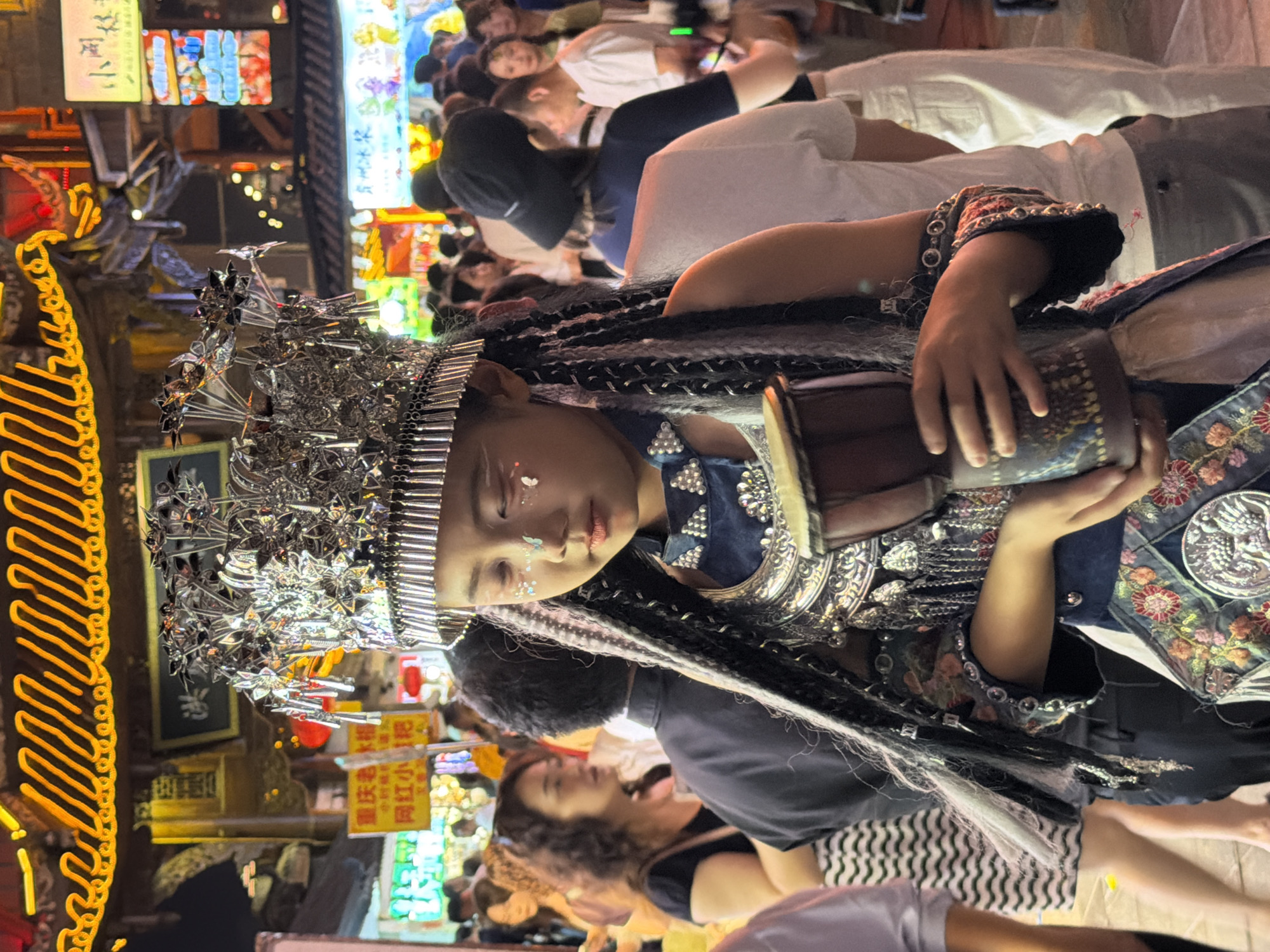 This media file captures a vibrant street scene at night, likely in the Yuzhong District of China, with a prominent young girl in the foreground. The girl is adorned in an elaborate, silver headdress and traditional-style clothing, featuring intricate embroidery and decorative elements. She holds a small drum in her hands, suggesting a performance or celebration. Her makeup includes sparkling embellishments on her face.
The background is a lively and colorful streetscape illuminated by numerous neon signs and glowing lights, indicative of a commercial or entertainment district after dark. Several people are visible in the background, some appearing to be spectators or participants in the event. The overall atmosphere seems festive and engaging.
The image is vertically oriented and the focus is primarily on the girl and her attire. The bright lights and busy street suggest a bustling urban environment. The time of day is clearly nighttime. The scene evokes a sense of cultural celebration or performance within a lively urban setting. Some visible text appears on signs in the background, though it is not clearly legible.