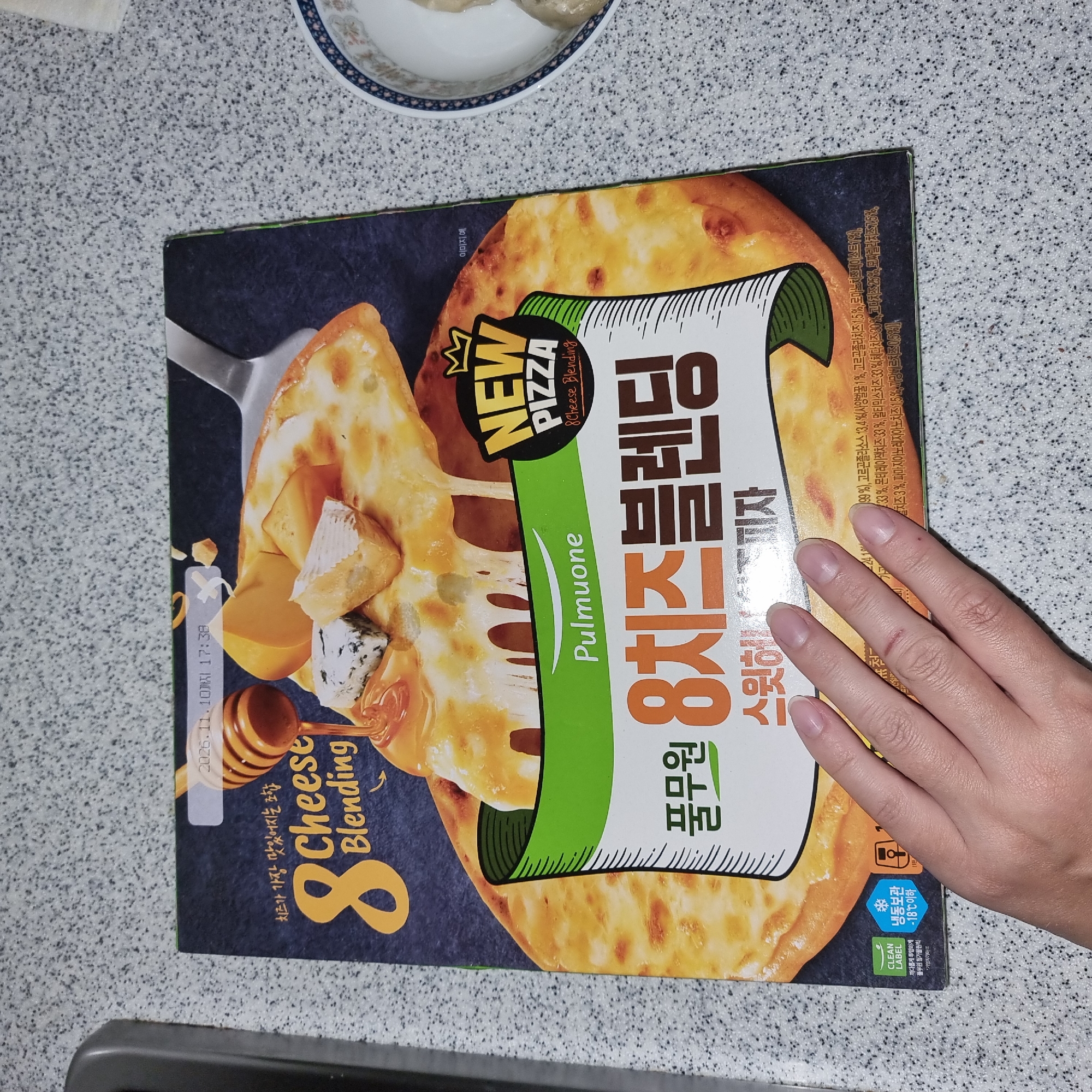 A person's hand is visible in the foreground of the image, holding down a box of Pulmuone brand 8 Cheese Blending pizza. The pizza box is depicted in the center of the frame, with an image of a cheesy pizza with honey drizzled on it. Text on the box indicates the product is "8 Cheese Blending" and the brand name "Pulmuone" is prominently displayed. Other text in Korean and English suggests it's a new pizza product with various cheese blends. A date of "2026.11.10" is visible, likely an expiration date. The background shows a speckled countertop. A bowl with what appears to be dumplings is visible to the right of the pizza box. The image appears to be taken indoors, likely in a kitchen or dining area. There is no visible indication of the specific location within Dunnae-myeon, South Korea.